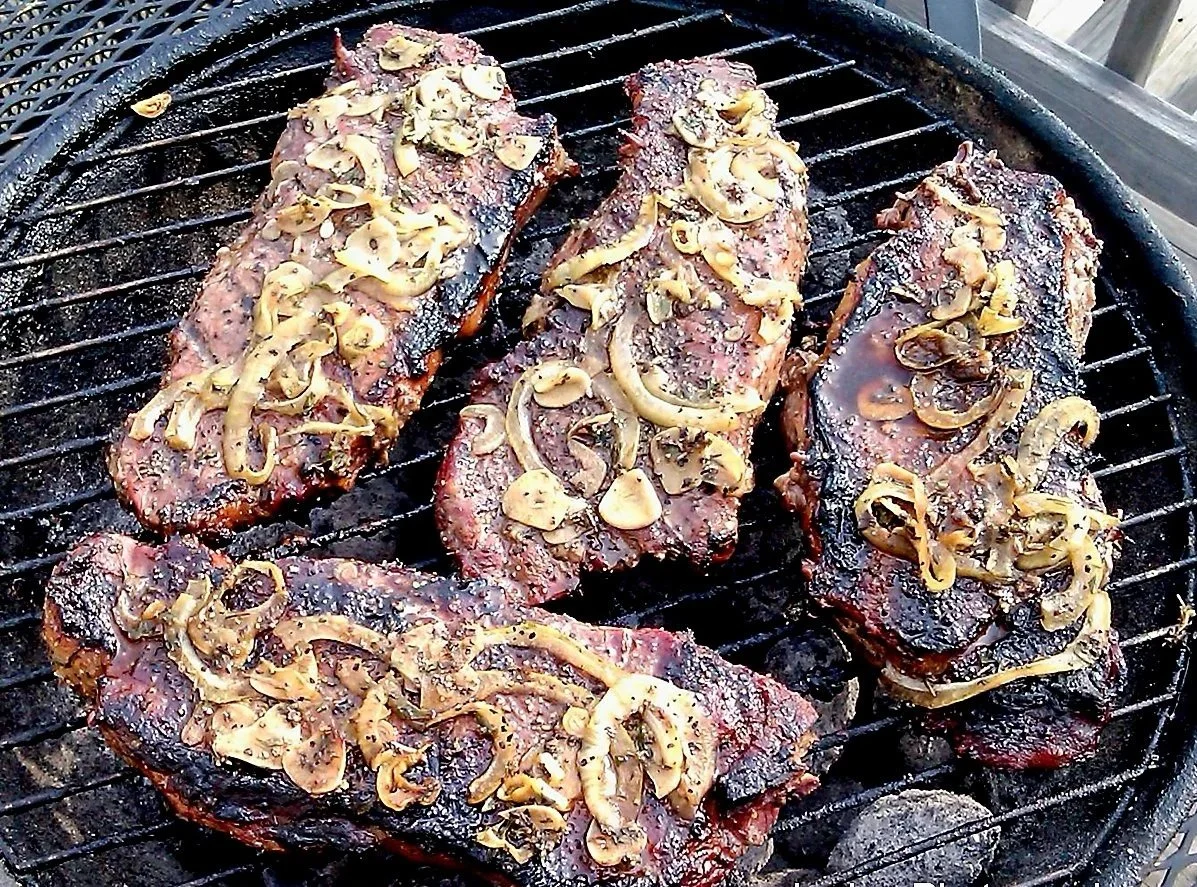 Meat steaks on a grill, topped with sliced  caramelized onions.