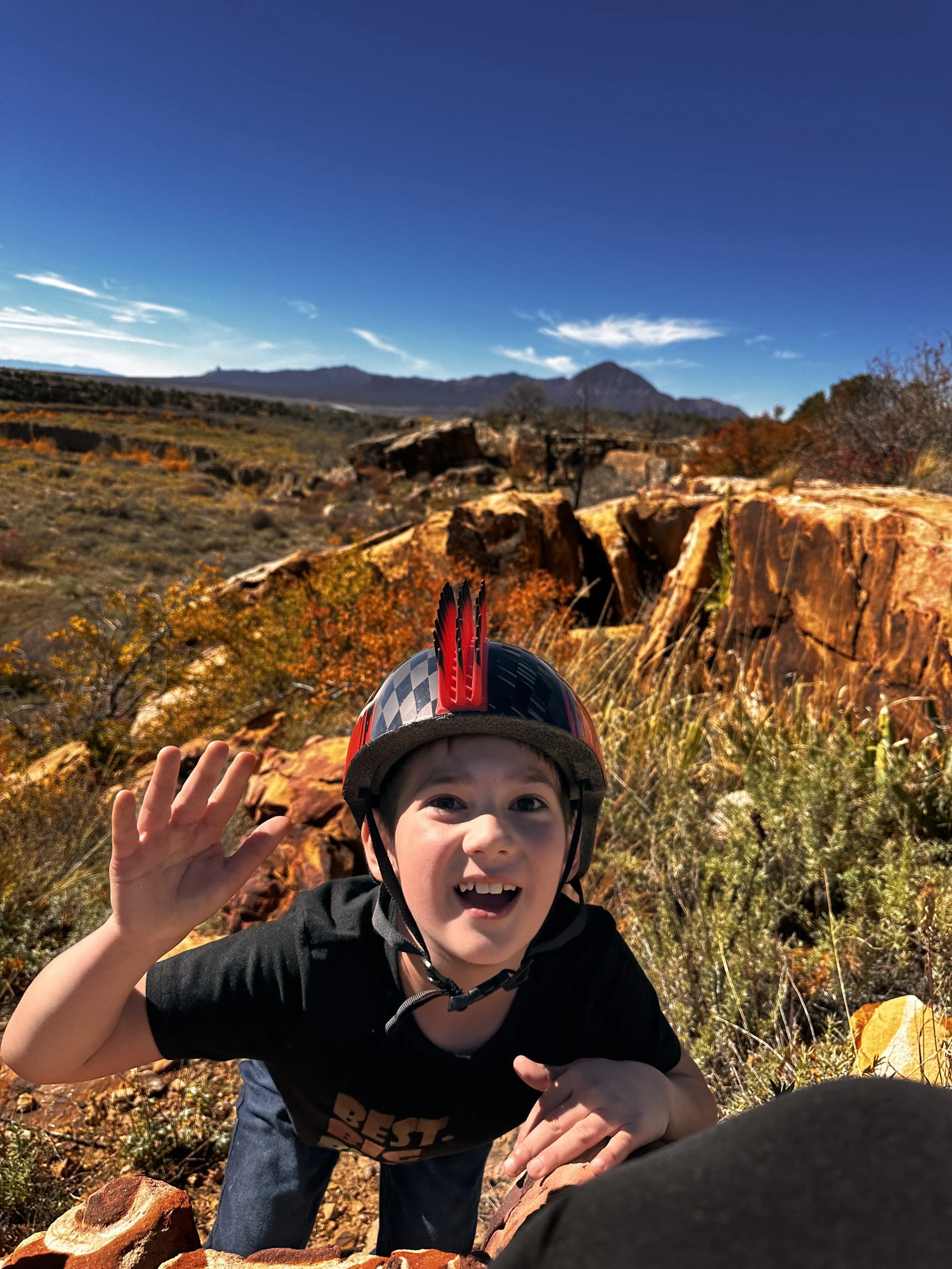 A young boy wearing a black helmet with red spikes, waving and smiling outdoors in a rocky, mountainous landscape with blue sky, distant mountains, and autumn foliage.