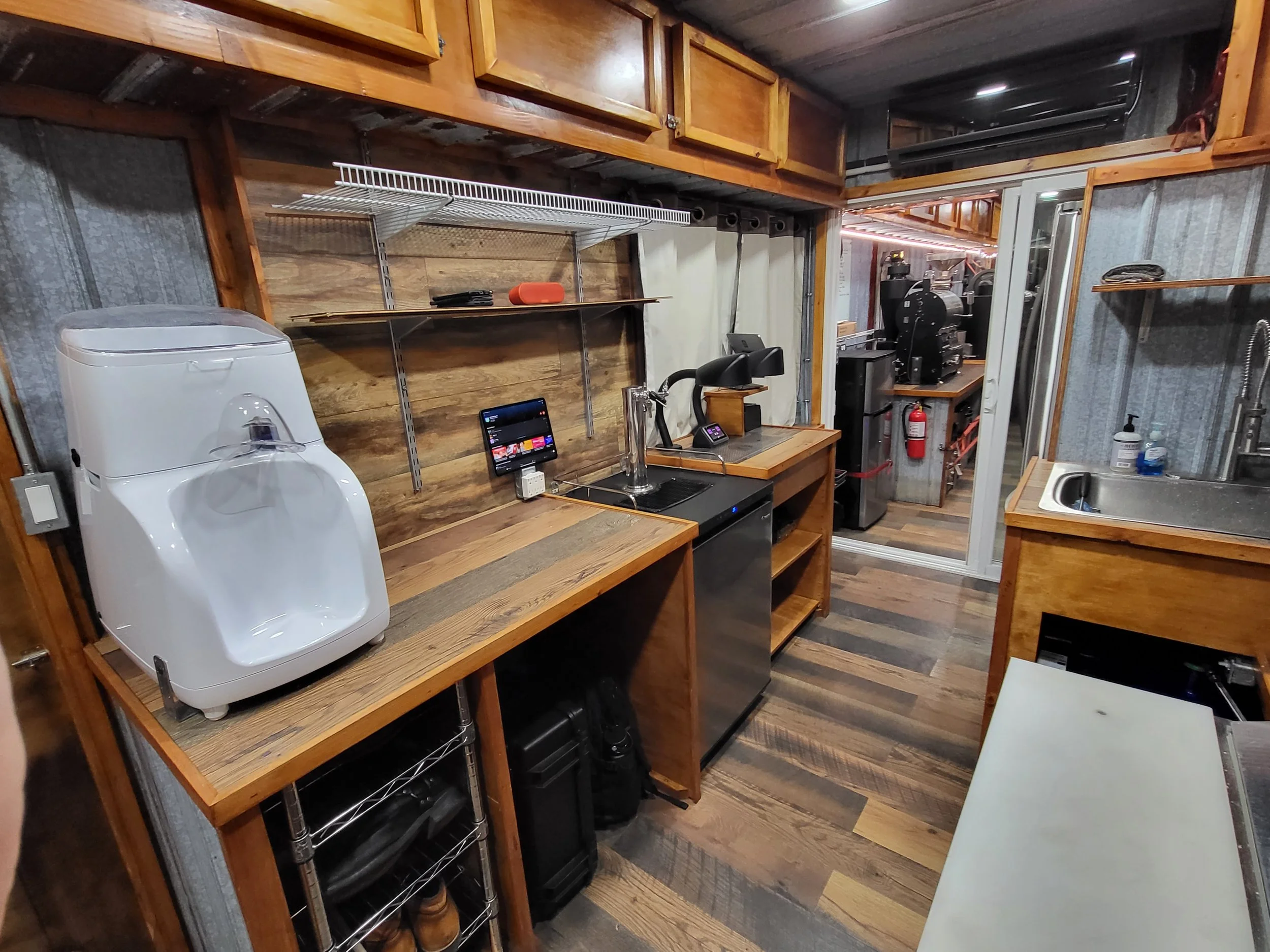A coffee shop workspace with a white coffee machine on a wooden counter, a small tablet or screen, a black coffee grinder, and various kitchen tools. The interior features wooden cabinets, shelves, and textured walls, with visible equipment and suppl