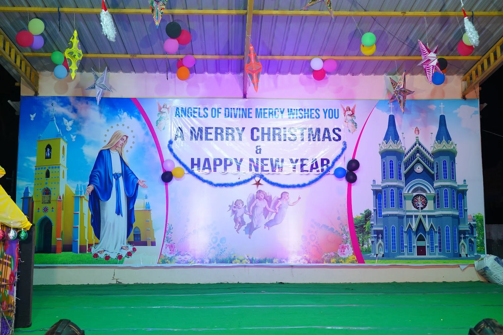 Colorful Christmas and New Year greeting banner with religious imagery, including a depiction of the Virgin Mary on the left and a church on the right, decorated with ornaments, balloons, and stars, with the message wishing a Merry Christmas and Happ