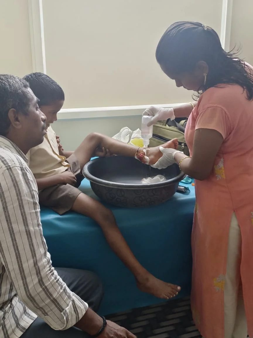 A woman in a coral outfit and gloves is tending to a young boy's arm in a medical setting. The boy is sitting on a bed, with his leg extended over a basin. A man is seated beside him, observing. The woman appears to be preparing to treat or bandage t