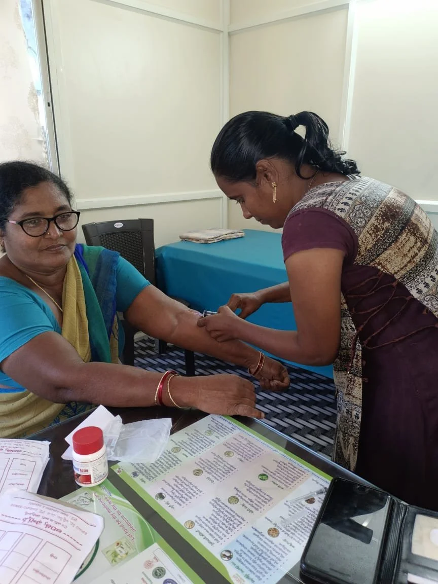 A woman sits at a table with her arm extended while a healthcare worker prepares to administer a shot. The table has medical supplies and informational pamphlets. The setting appears to be a clinic or health center.