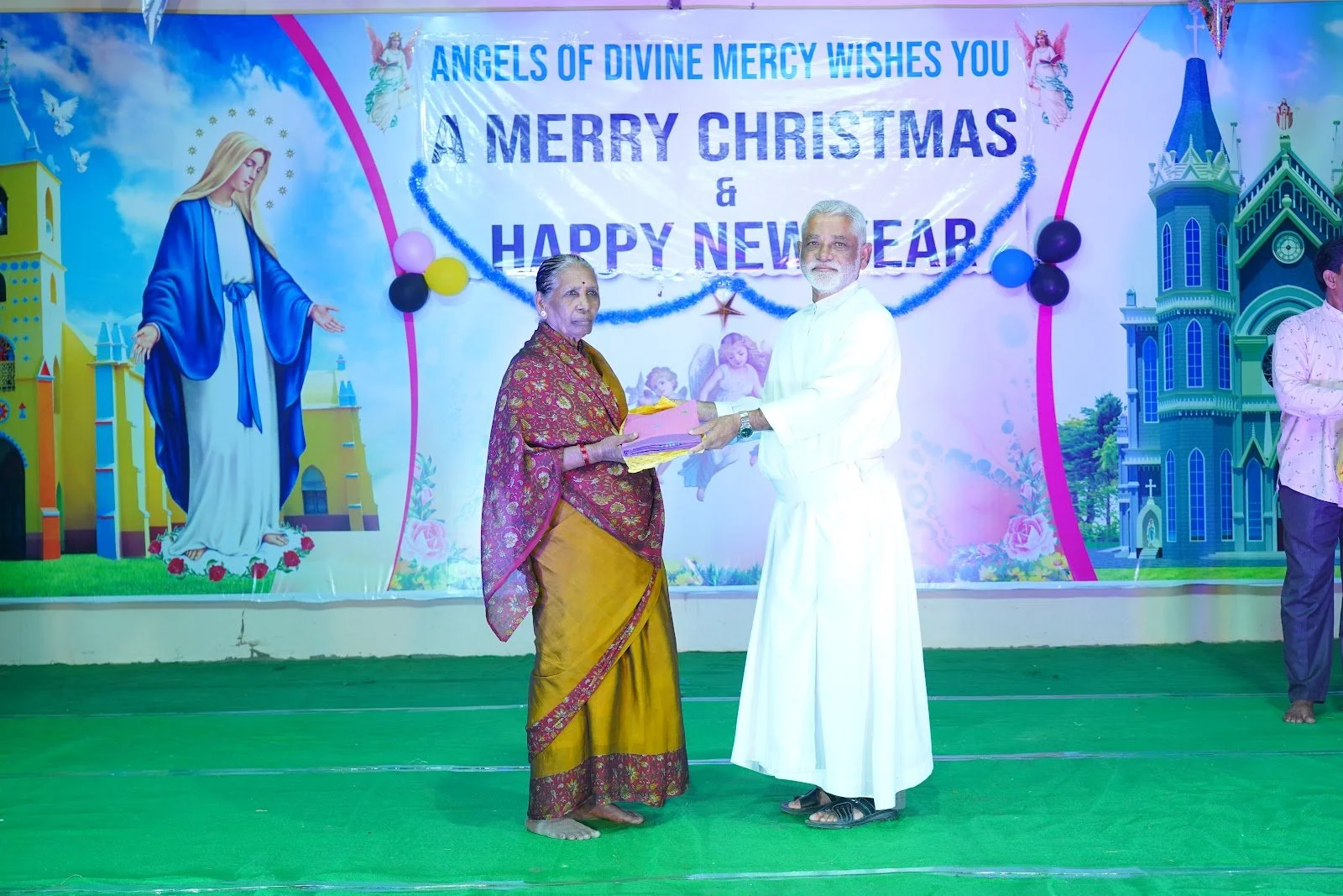 A woman in traditional saree receiving a gift from a man dressed in white on a stage decorated for Christmas and New Year celebration, with a backdrop featuring religious and festive imagery, including Jesus and angels.