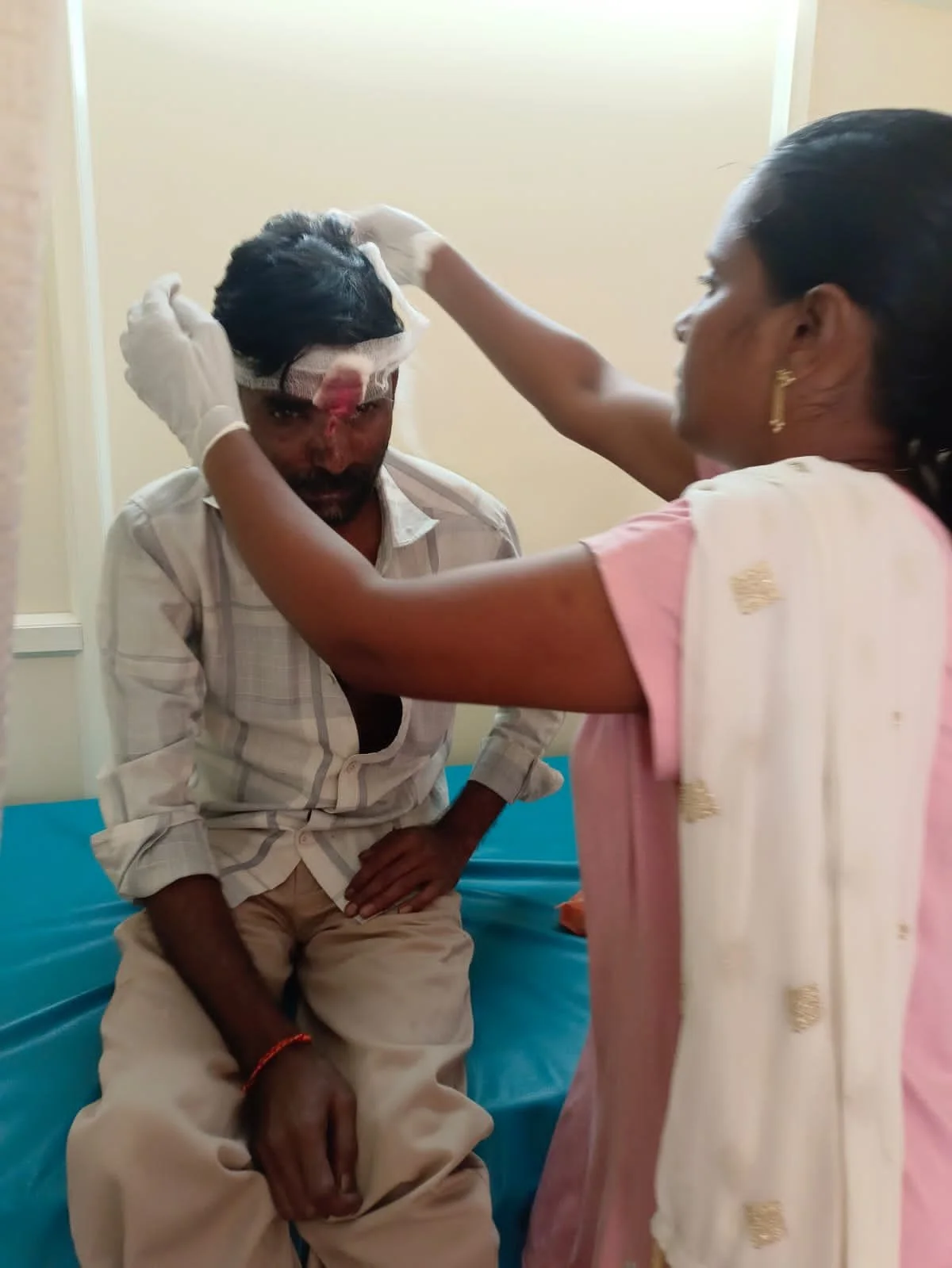 A woman dressed in pink is placing a bandage on the head of a man who is sitting on a medical bed, in what appears to be a healthcare setting.