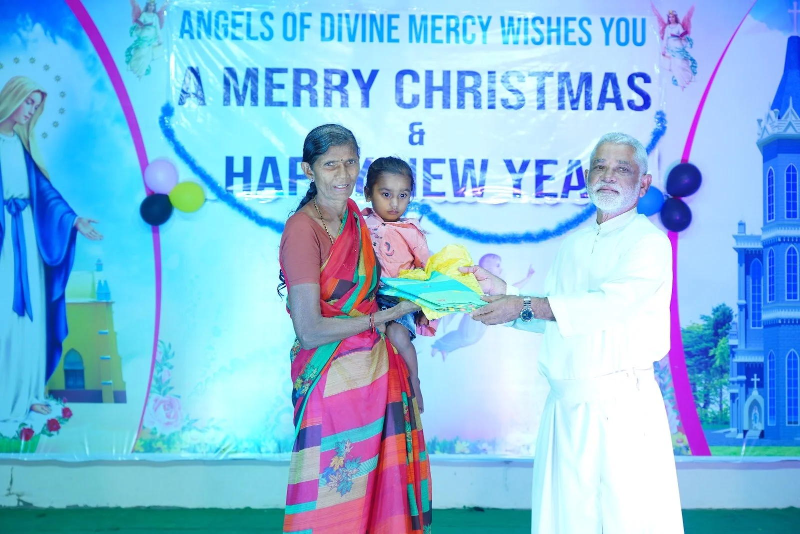 A man is giving a gift to a woman holding a girl at a Christmas and New Year celebration event. The background features a festive banner with religious imagery and text wishing Merry Christmas and Happy New Year.