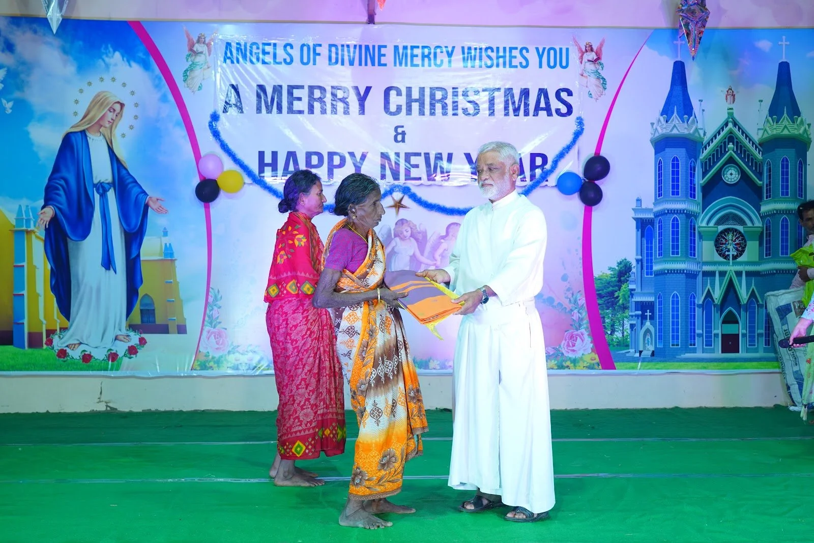 A man in white giving a folded cloth to an elderly woman in a patterned saree, with two other women standing nearby, in front of a colorful Christmas and New Year greeting banner with religious imagery and architecture.