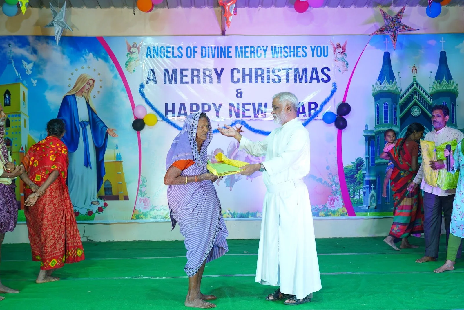 A church event where an elderly woman receives a gift from a man in white robes, with a colorful backdrop featuring religious imagery, including Jesus and a church, and a greeting that says 'Angels of Divine Mercy Wishes You a Merry Christmas & Happy