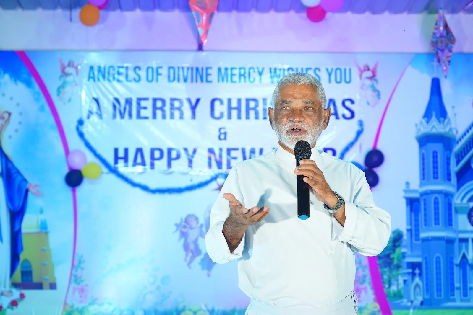 An elderly man with white hair and beard holding a microphone, speaking at a Christmas and New Year celebration. Behind him is a decorated backdrop with Christmas-themed illustrations and the words 'Angels of Divine Mercy Witches You, A Merry Christm