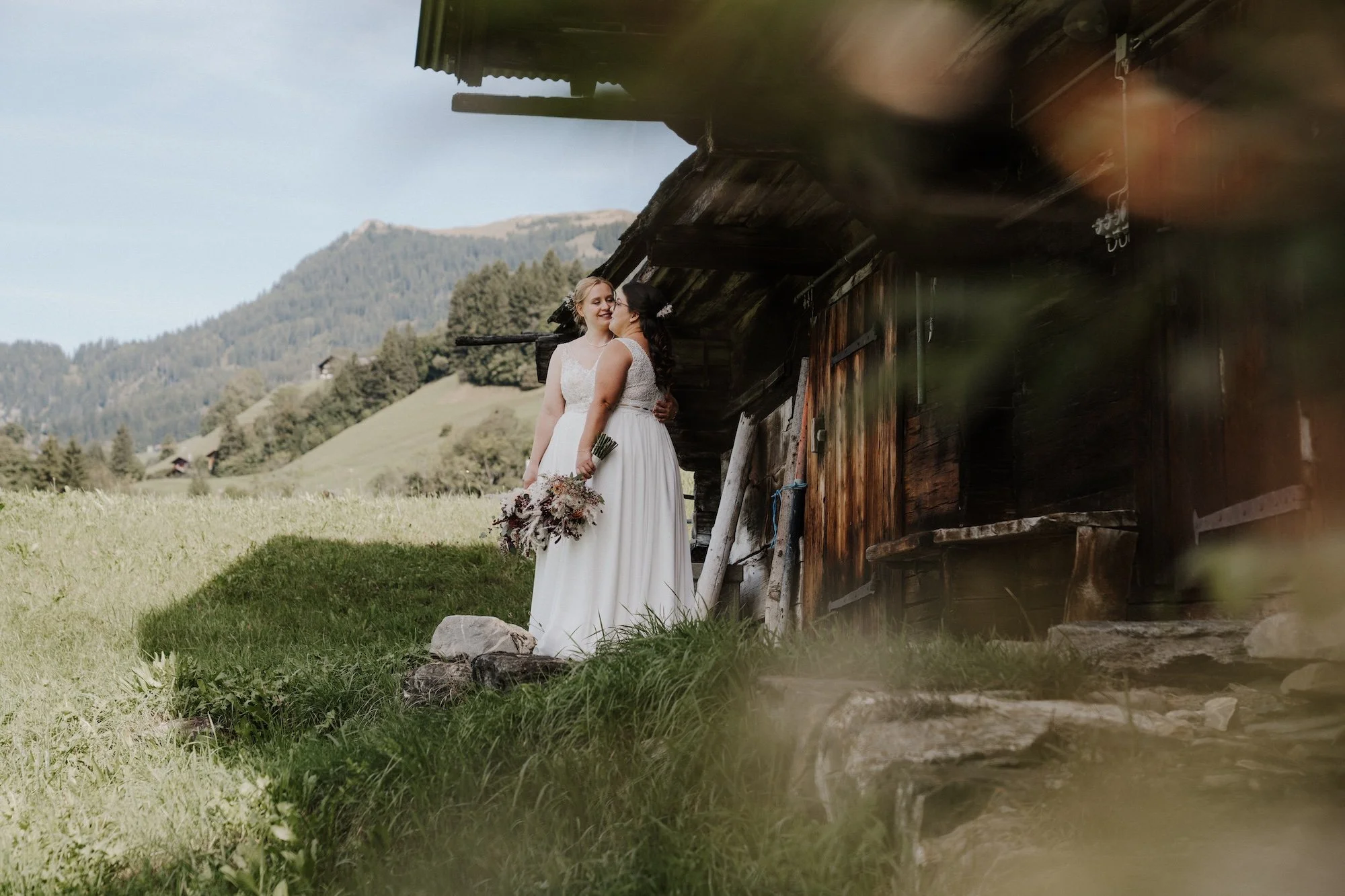 Nadine und Svenja vor einem alten Holzhaus auf dem Hasliberg – eine queere Hochzeit in den Schweizer Bergen.