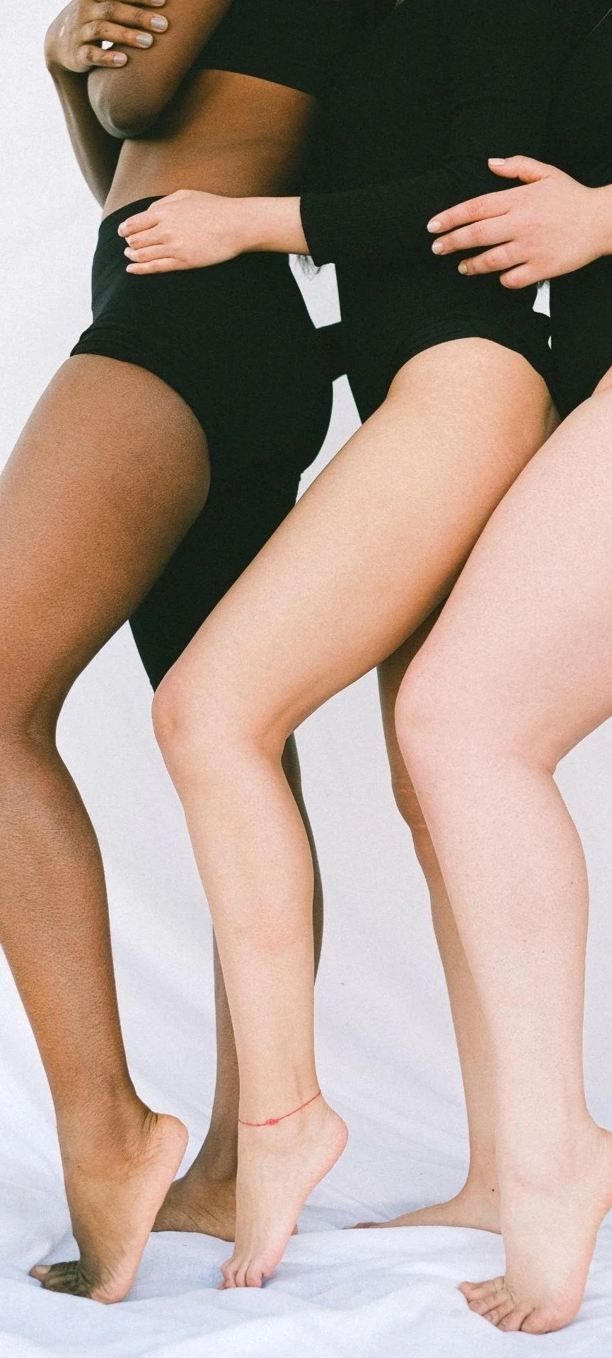 Drei Frauen stehen eng beieinander, nur ihre Beine und Arme sind sichtbar, mit unterschiedlichen Hauttönen, auf einem weißen Hintergrund.