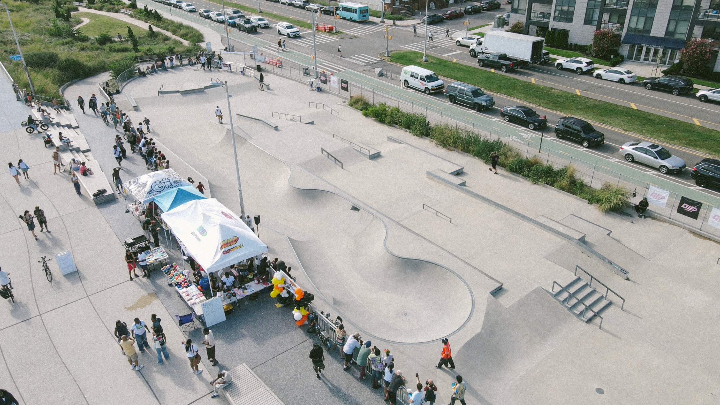 People gathering at an outdoor skatepark with tents and booths, cars parked along nearby streets, and urban buildings visible in the background.