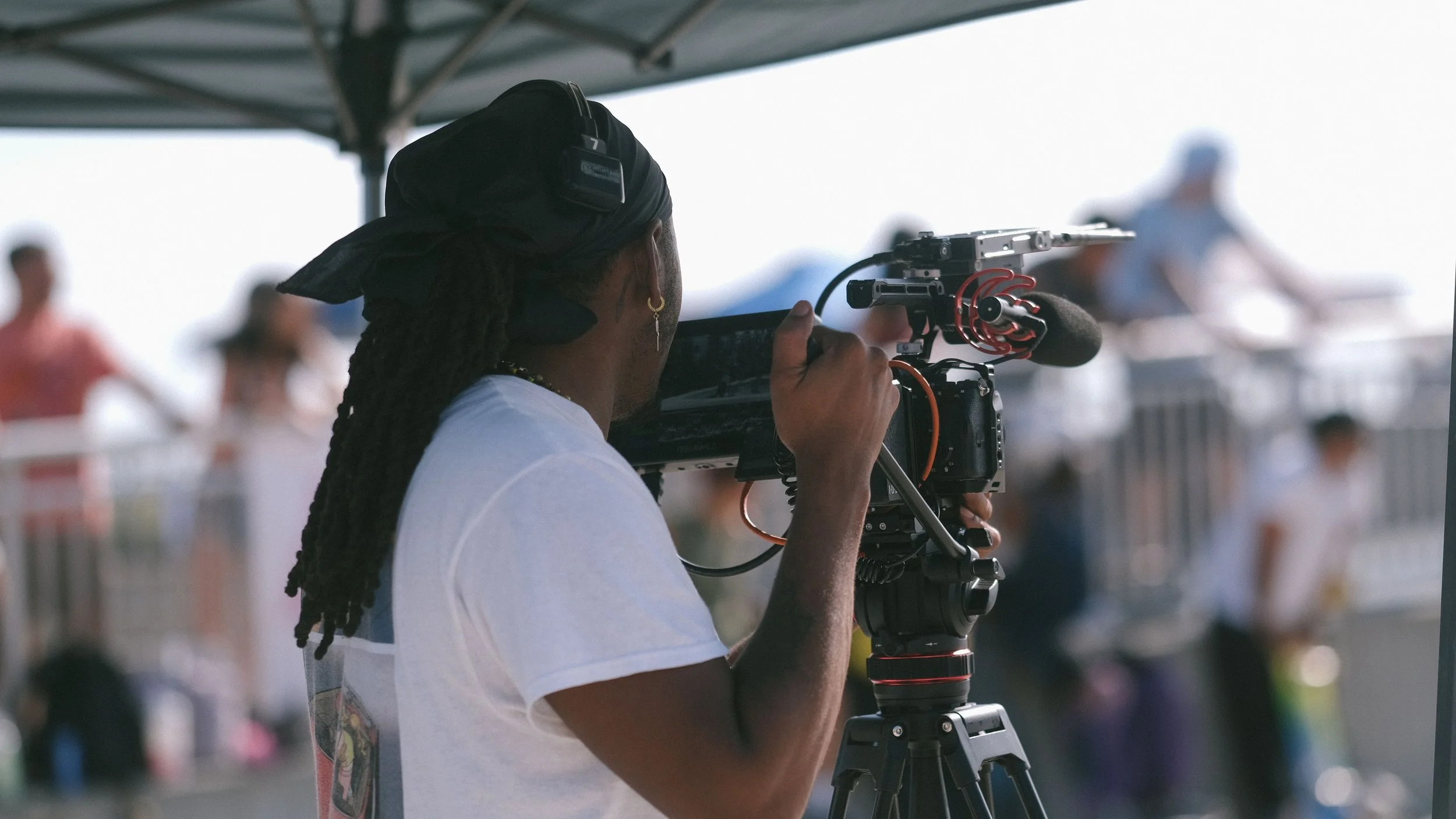 A person with long dreadlocks and a black head covering operating a professional video camera on a tripod at an outdoor event.