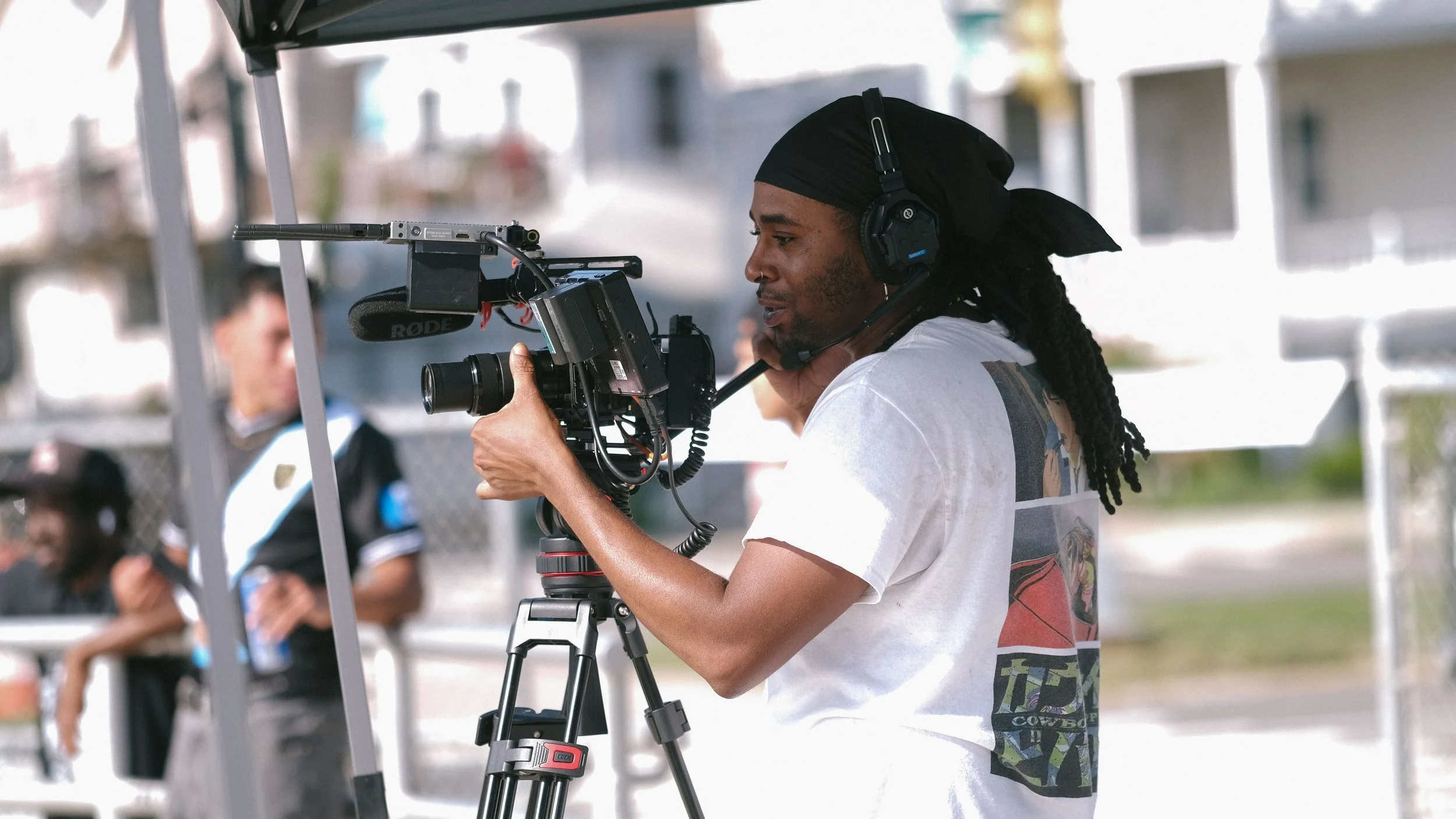 A person, possibly a videographer, wearing a black headscarf and headphones, operating a professional video camera mounted on a tripod outdoors.
