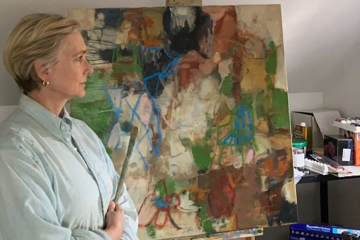 A woman with curly gray hair wearing a black hat, pink glasses, a black blazer, and yellow plaid pants, standing and speaking in front of an abstract painting in an art gallery.