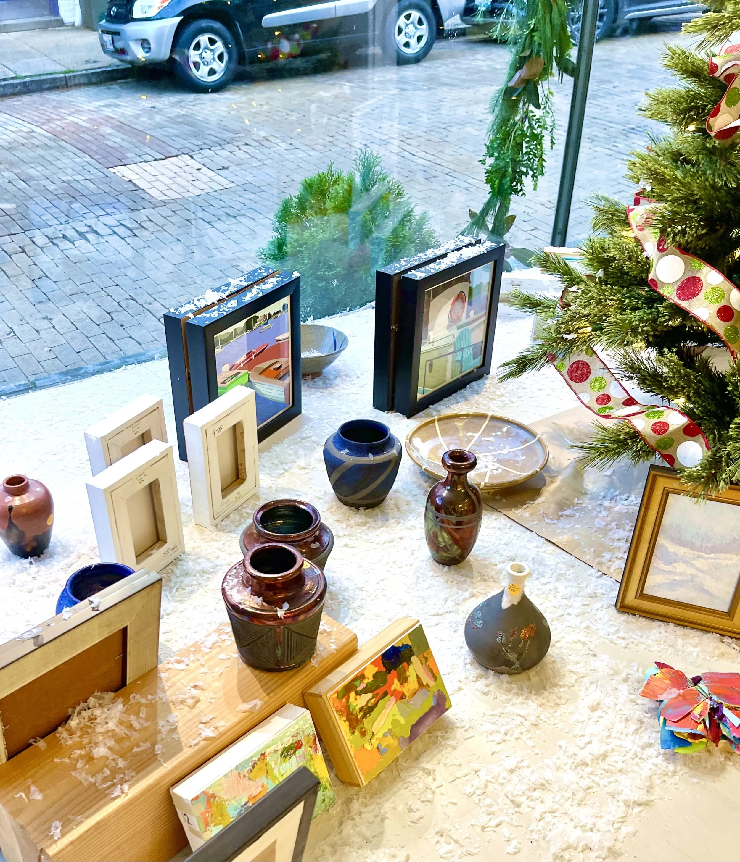 Display window with various art pieces and pottery, Christmas tree with ribbon, snow on surface, and cars parked outside street.