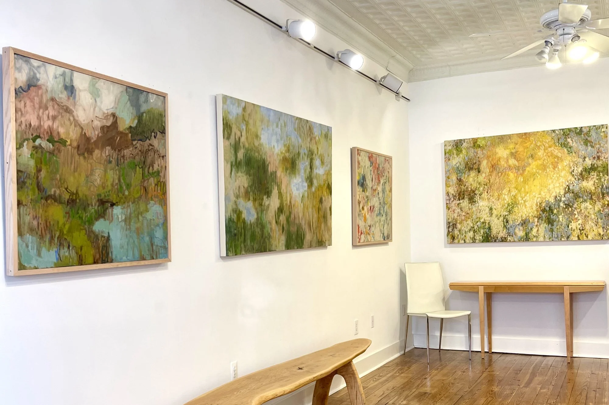 An art gallery with four abstract landscape paintings on white walls, a wooden bench, a white chair, and a wooden table on a hardwood floor, with ceiling lights and a ceiling fan.