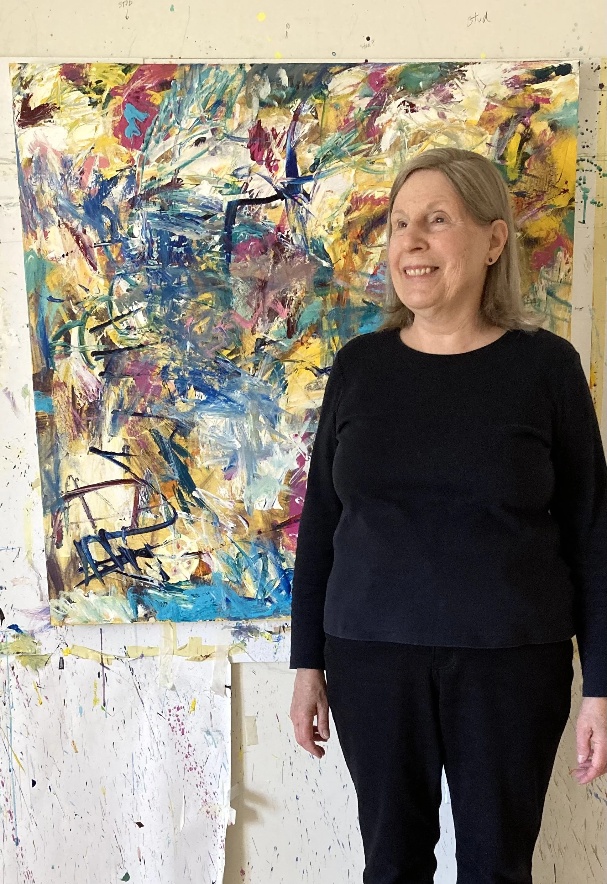 A woman with curly gray hair wearing a black hat, pink glasses, a black blazer, and yellow plaid pants, standing and speaking in front of an abstract painting in an art gallery.