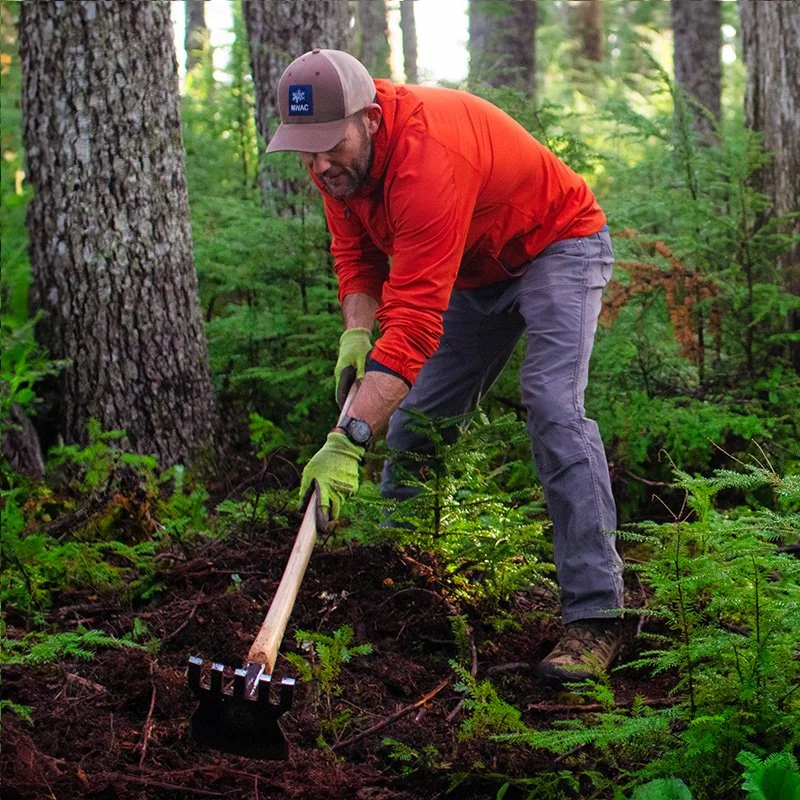 A man in an orange jacket and cap using a mattock in a forested area.