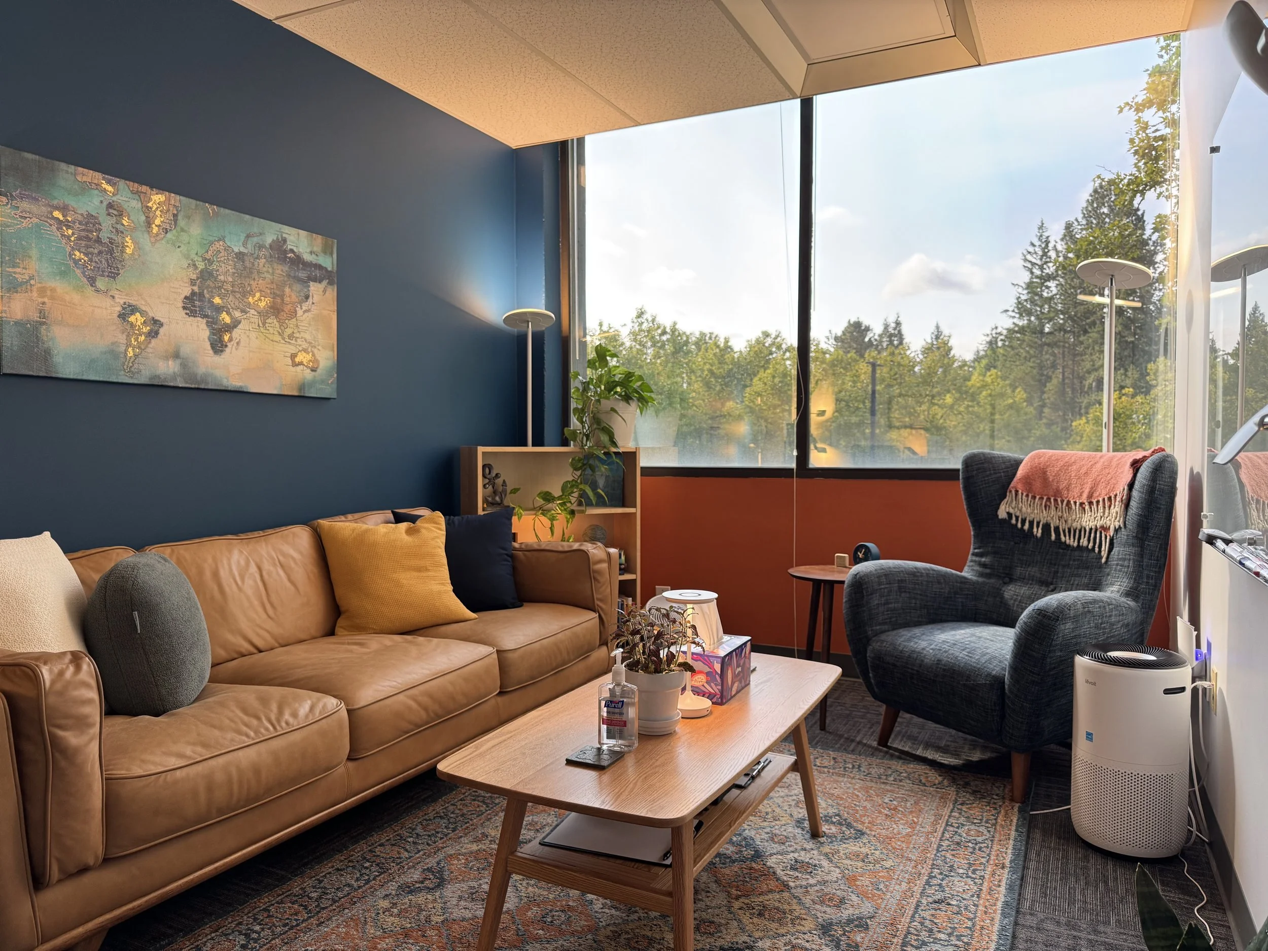 Cozy office lounge with a tan leather sofa, a black armchair with a pink throw, a wooden coffee table, large windows with a view of trees, a world map wall art, plants, and a white air purifier.