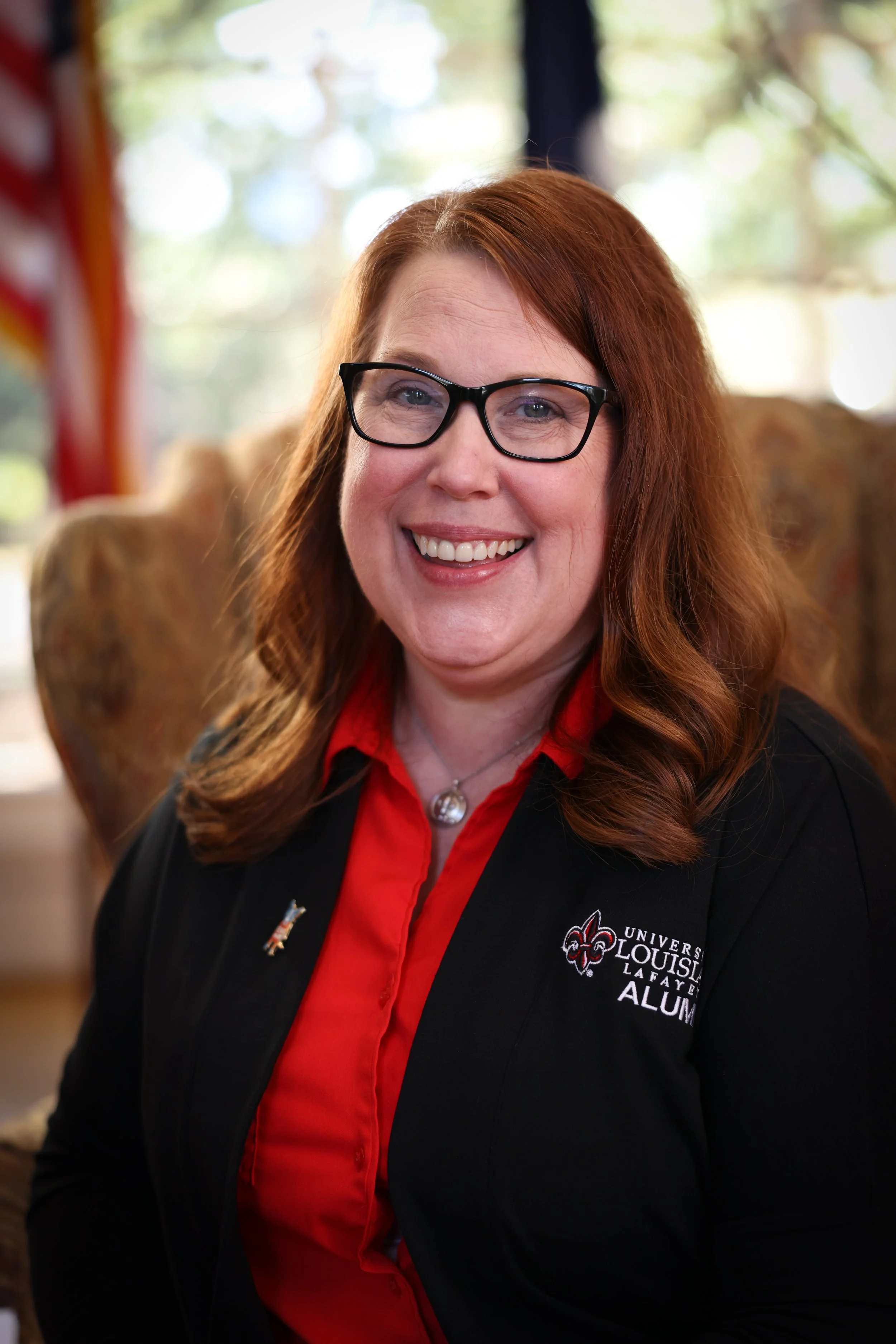 A woman with red hair, glasses, and a smile, wearing a black blazer and a red shirt with University of Louisiana at Lafayette Alumni logo, sitting indoors with blurred background of trees and sunlight.