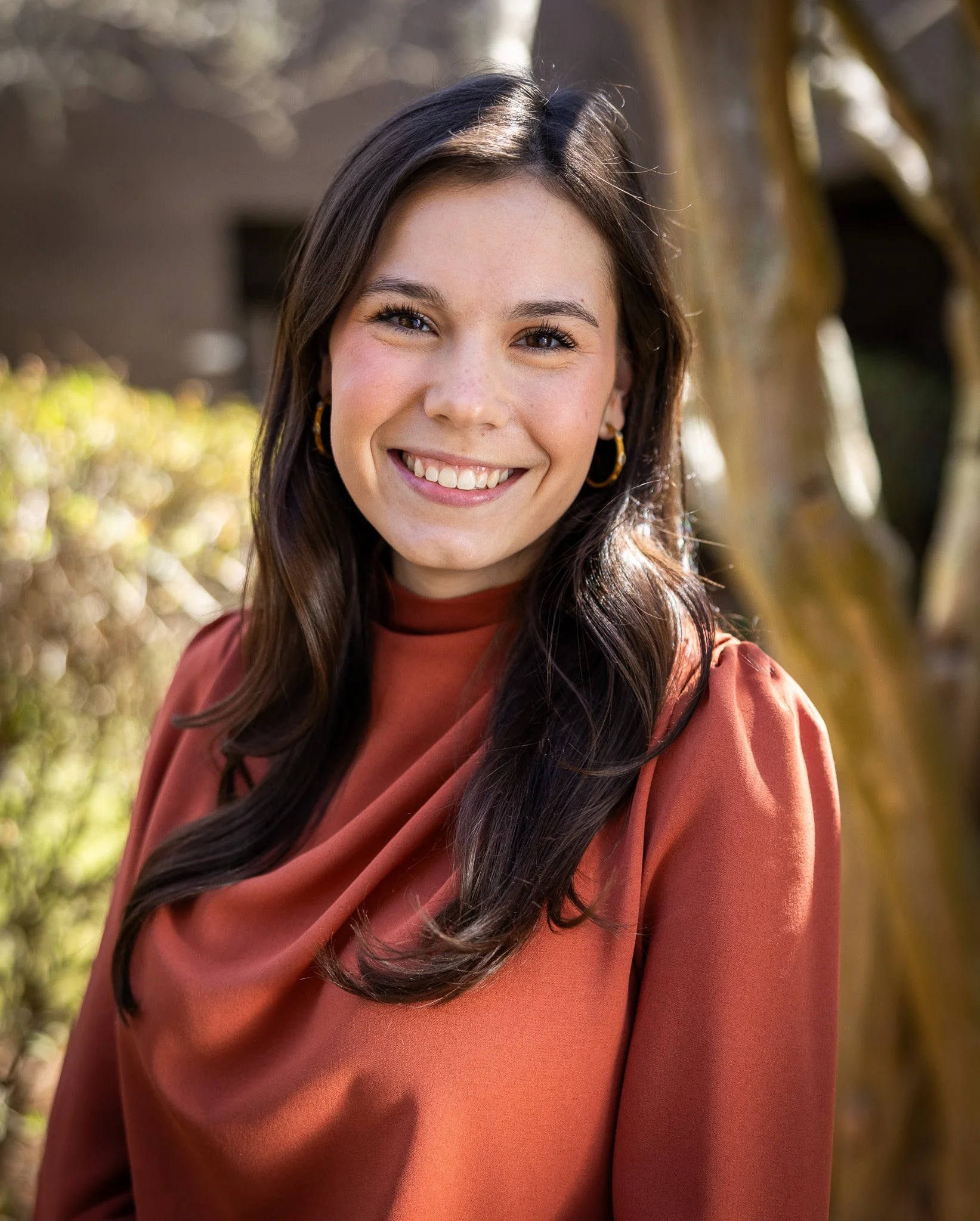 Close-up of a young woman with long dark hair, smiling, wearing gold hoop earrings and a rust-colored top, outdoors with blurred trees in the background.