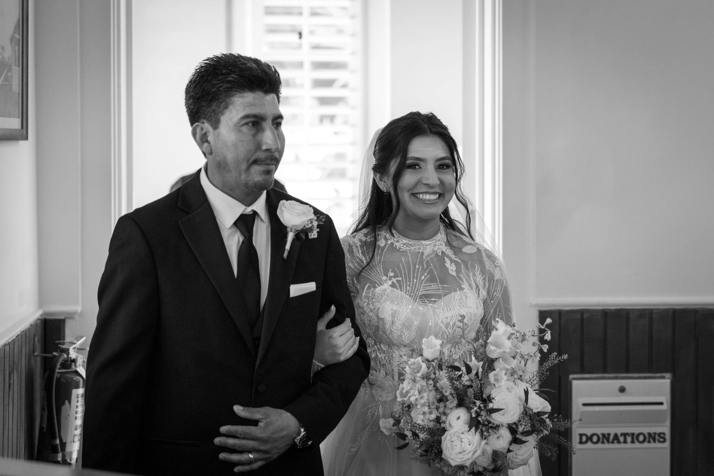 A black and white photo of a bride and groom standing together indoors. The bride is holding a bouquet of flowers, and the groom has a boutonniere on his suit. They are smiling and holding hands, participating in a wedding ceremony.