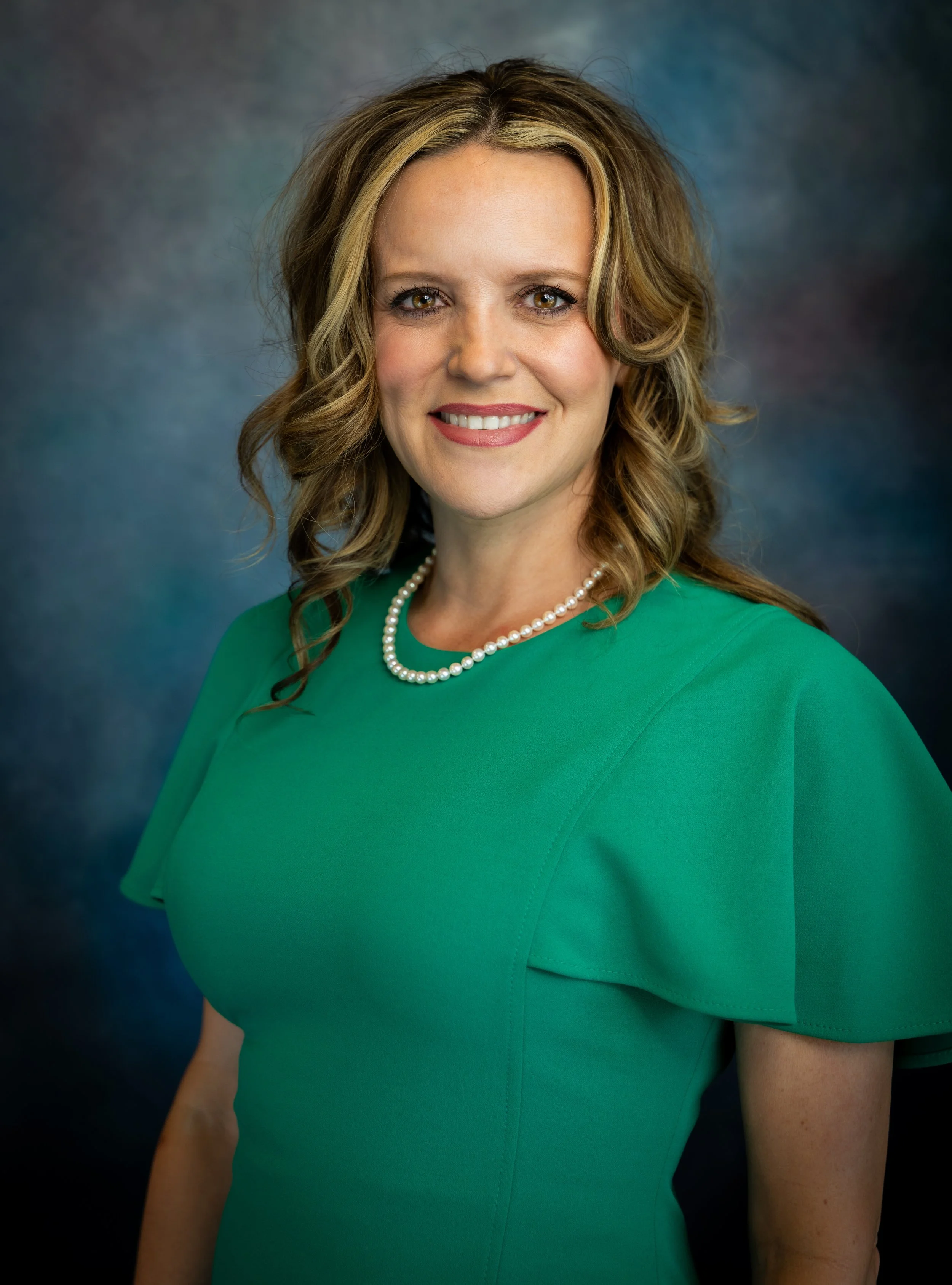 A smiling woman with light brown, wavy hair wearing a green dress and a pearl necklace, against a dark blue and green background.