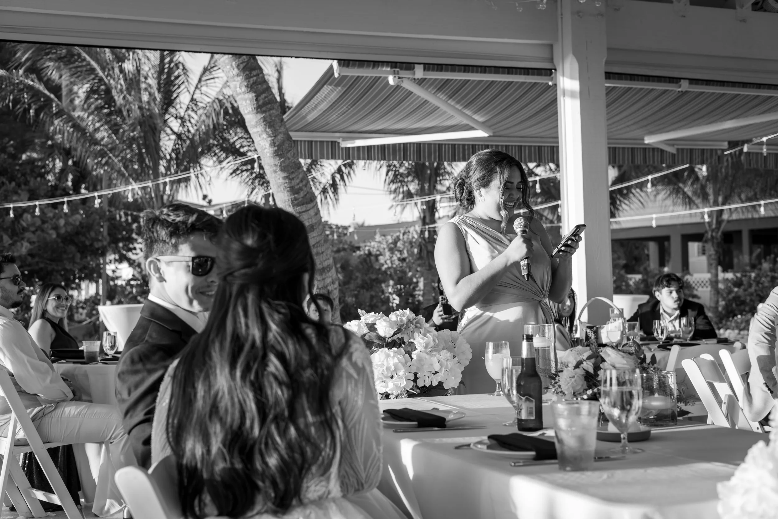 A woman in a dress giving a speech at an outdoor wedding reception, surrounded by seated guests, with a backdrop of palm trees and a partial striped canopy.