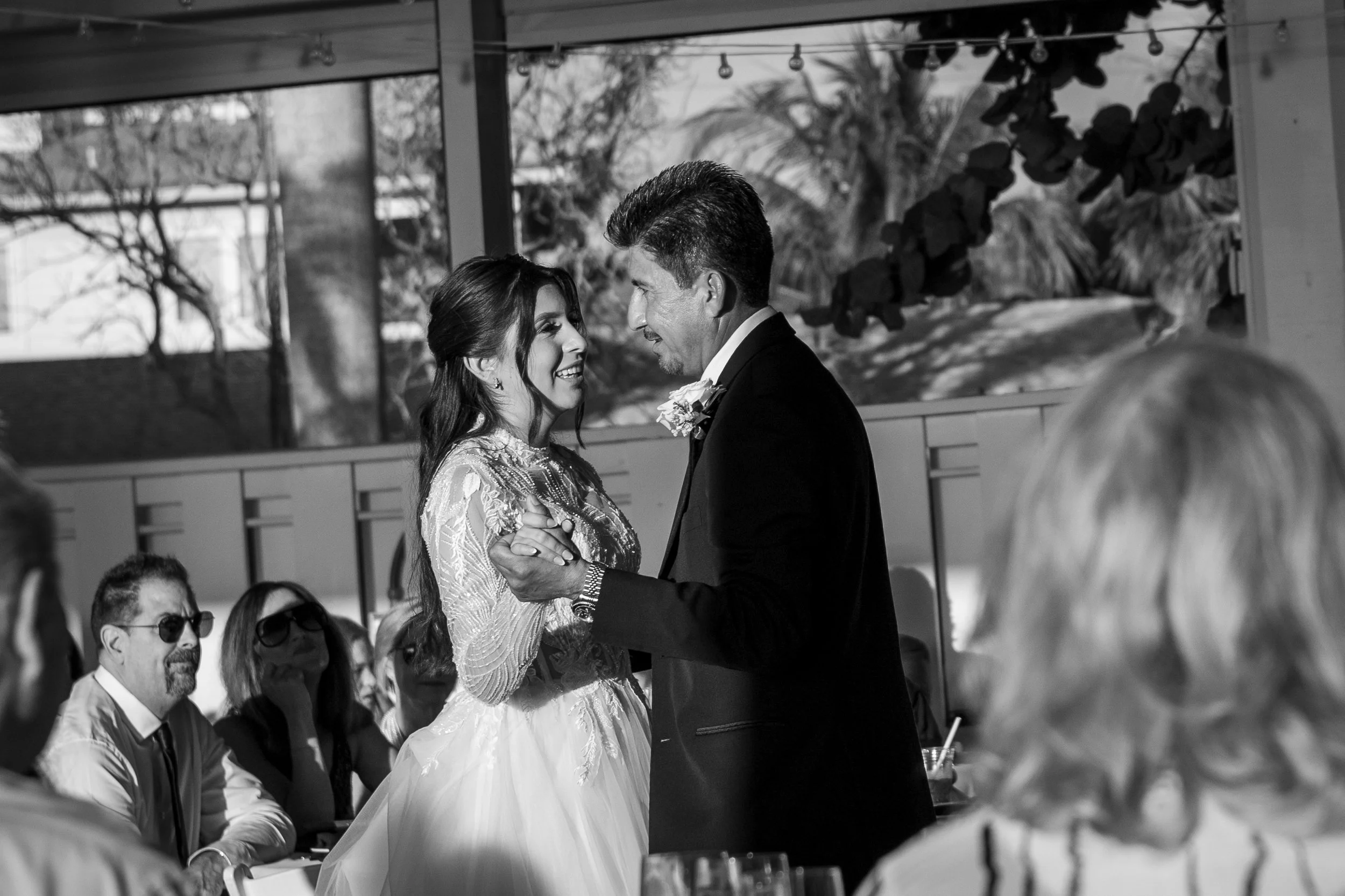 A black and white photo of a bride and groom dancing at their wedding reception, surrounded by seated guests, with trees visible outside through windows.