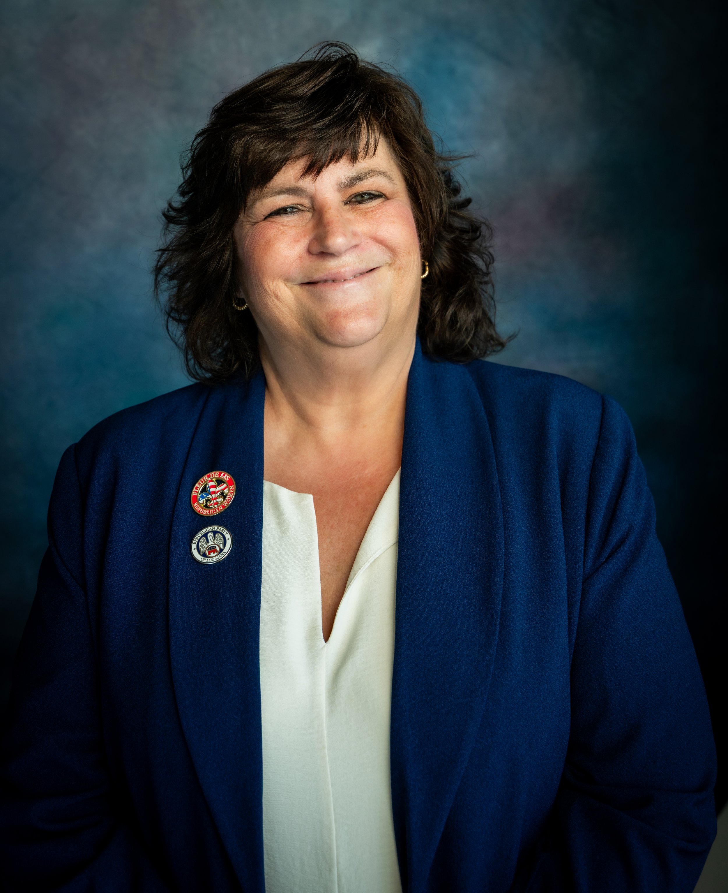 A woman with dark brown hair smiling, wearing a blue jacket with pins and a white blouse against a blue and green gradient background.