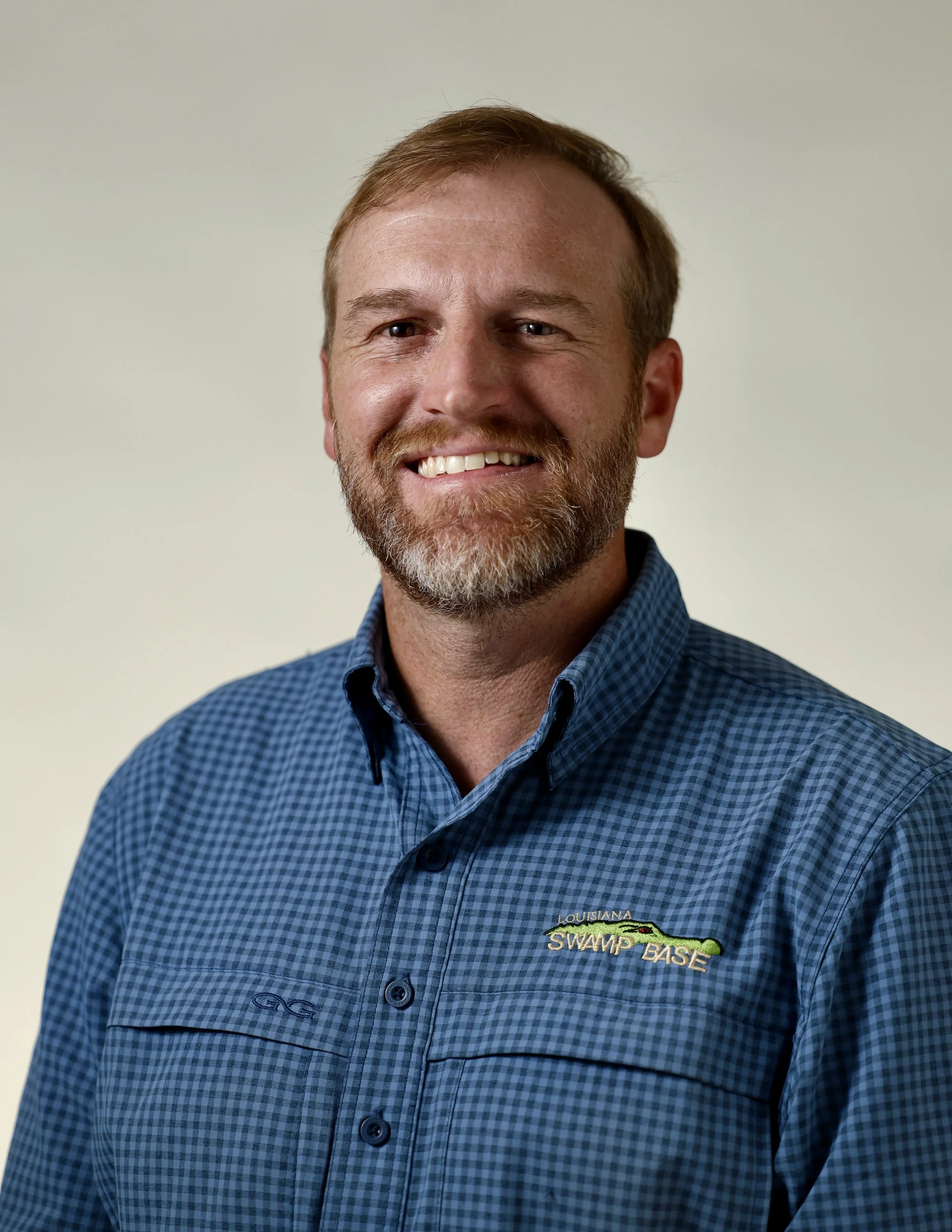 Close-up image of a smiling man wearing a blue checkered shirt with Louisiana Swamp Base embroidery.