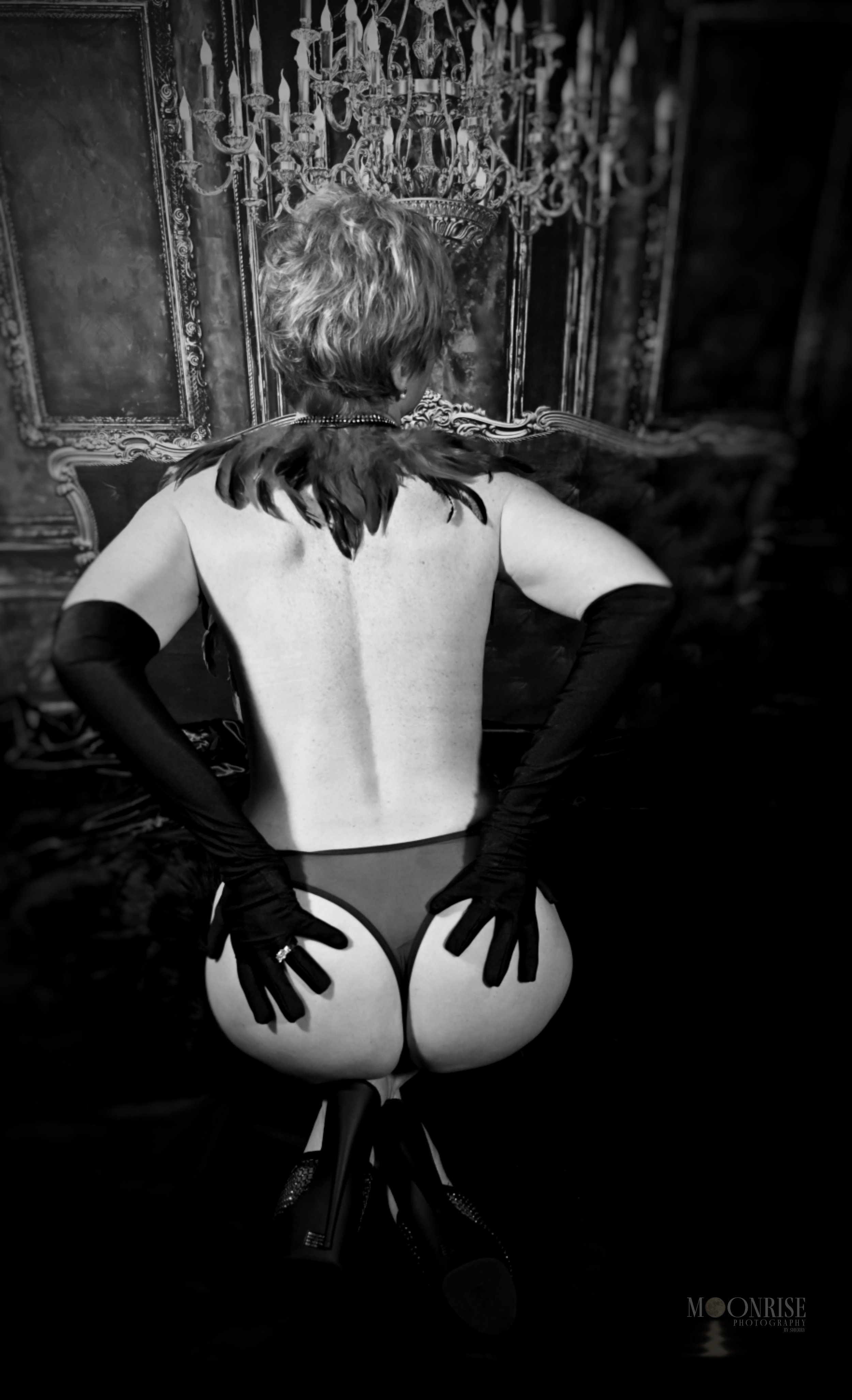Black and white photo of an individual sitting on a chair, facing away, wearing black lingerie, gloves, and a feather accessory under a chandelier.