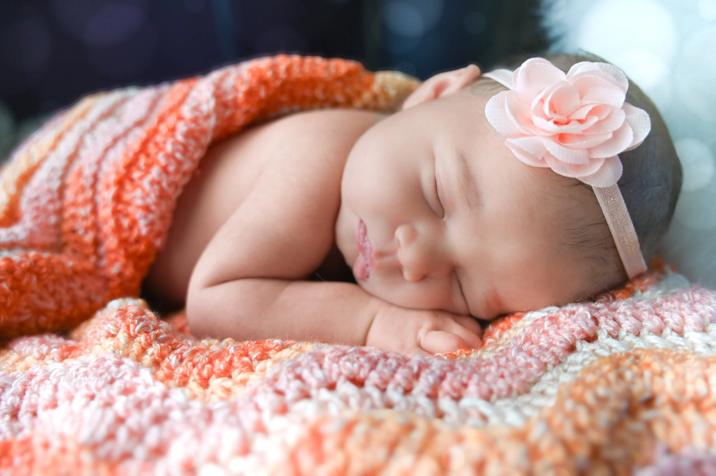 Newborn baby sleeping with a flower headband, wrapped in an orange and pink knitted blanket.