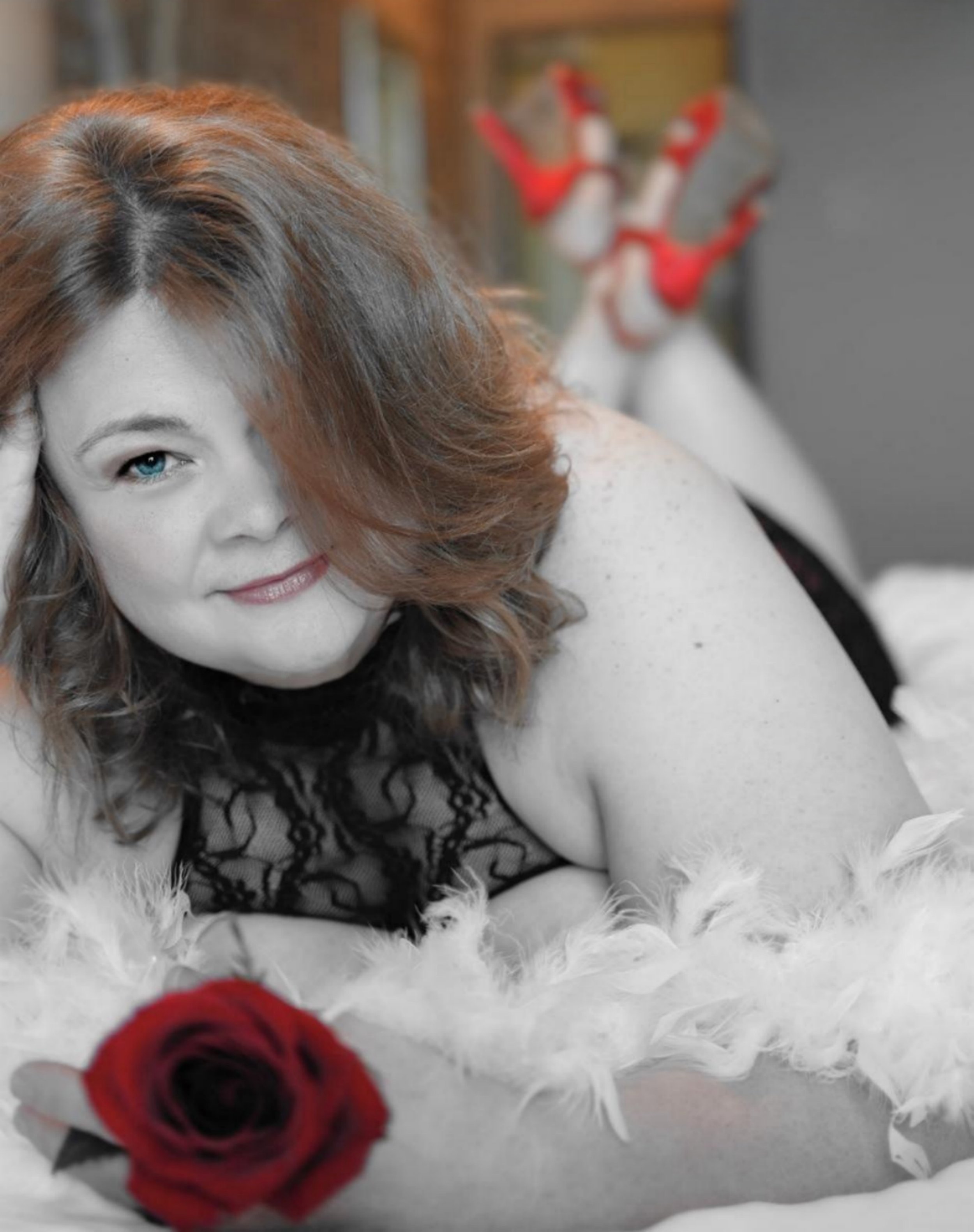 Woman lying on bed holding a red rose, wearing red high heels and lace top, with a white feather boa in the foreground.