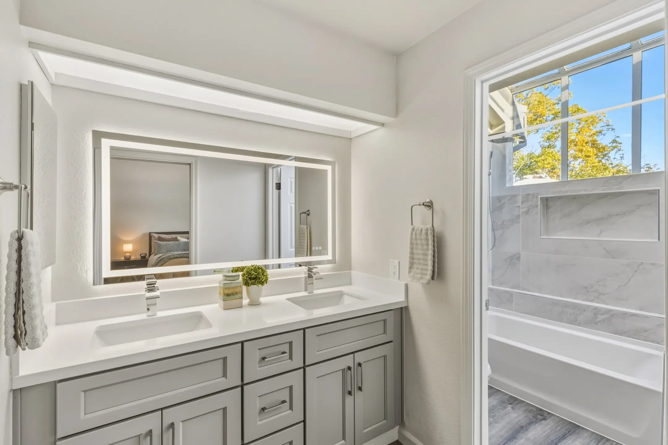 Modern white bathroom with double vanity, large illuminated mirror, and separate shower with marble walls and large window showing trees outside.