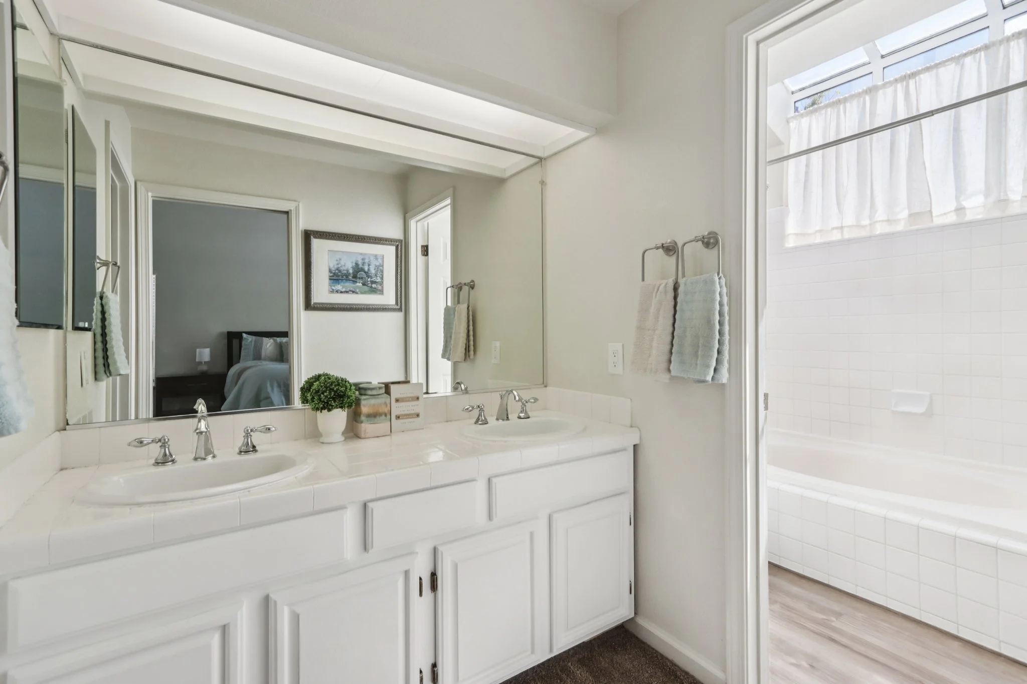 Bathroom with dual white sinks, large mirror, towels on towel rack, and a separate room with a bathtub and window with white curtains.
