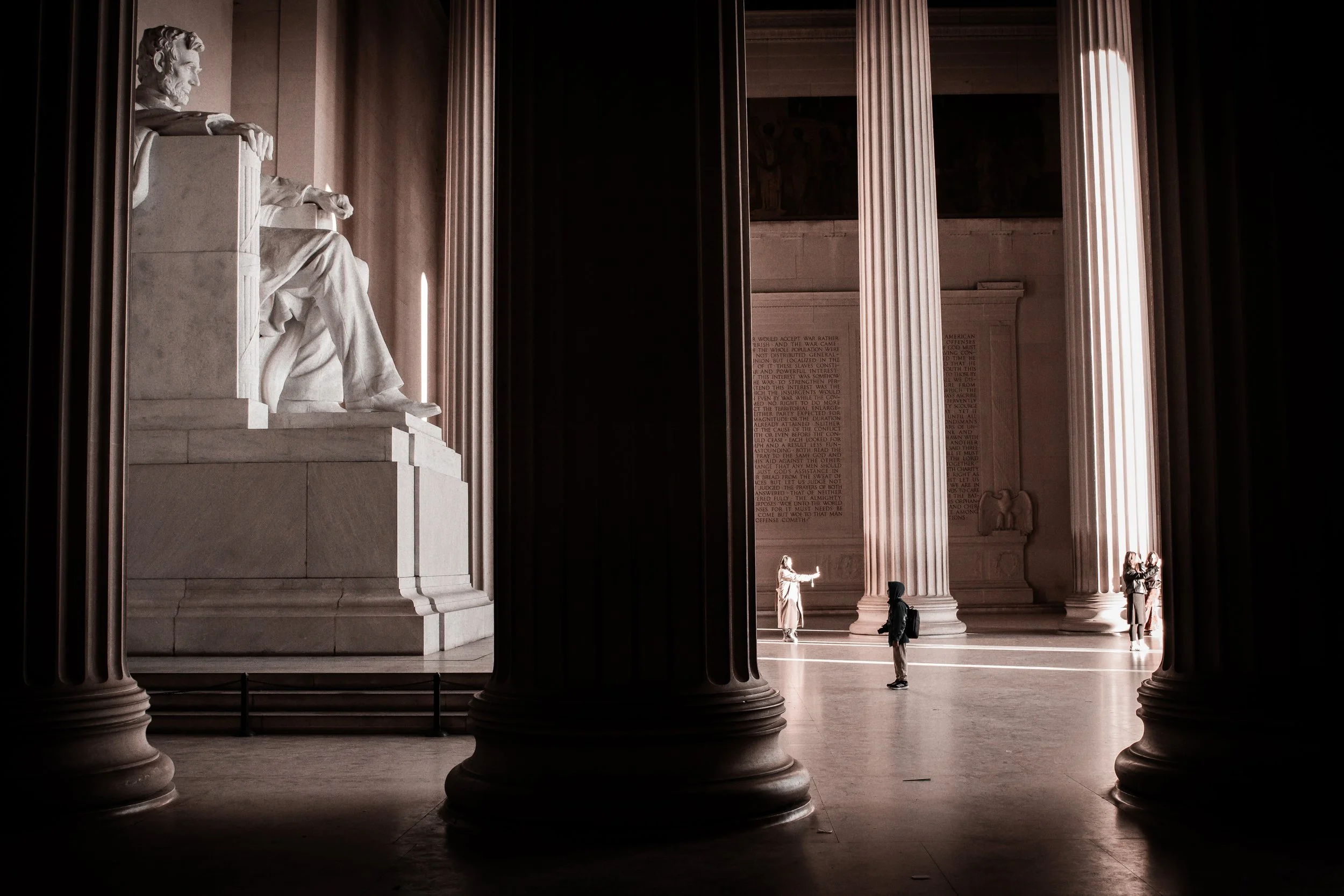 Interior view of the Lincoln Memorial with tall columns and a large seated statue of Abraham Lincoln. Two visitors are near the columns, and a large inscribed wall is visible in the background.