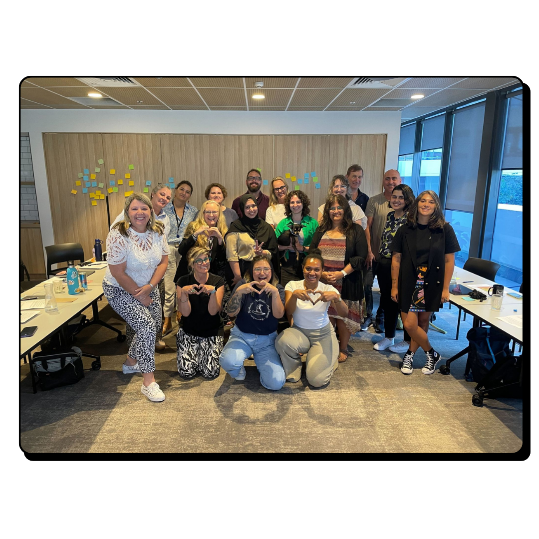 Group of diverse people smiling and posing for a photo in a modern office meeting room with sticky notes on the wall.