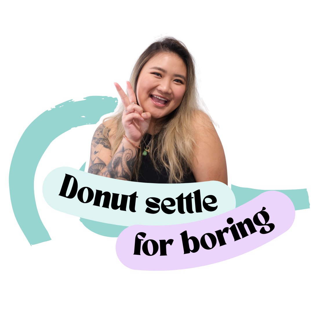 A young woman with long blonde hair, tattoos on her arm, smiling, making a peace sign with her hand, beside a sign that says 'Donut settle for boring'