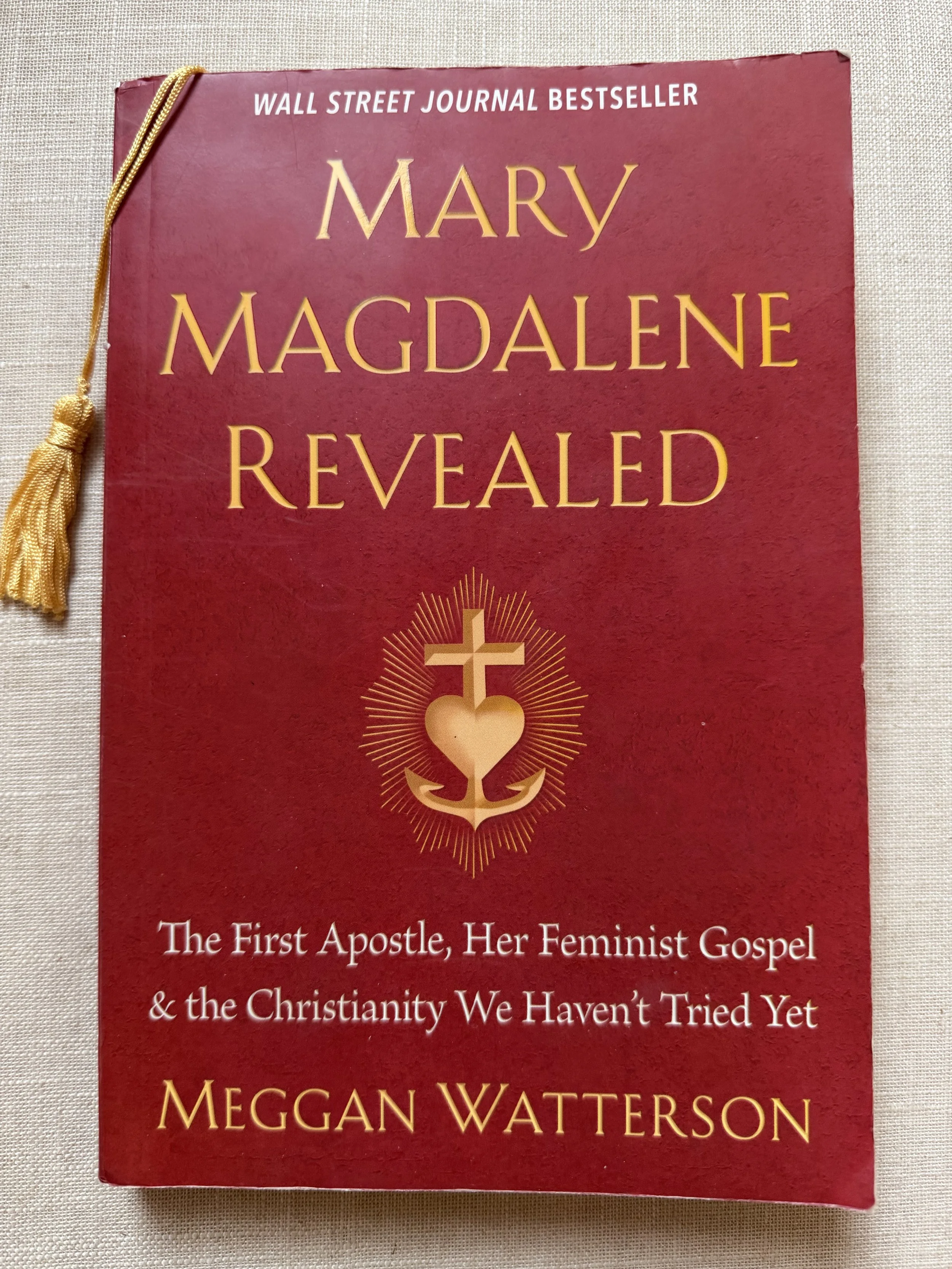 Red book cover of Mary Magdalene Revealed by Meggan Watterson, featuring a gold heart-and-anchor symbol and a tassel bookmark.