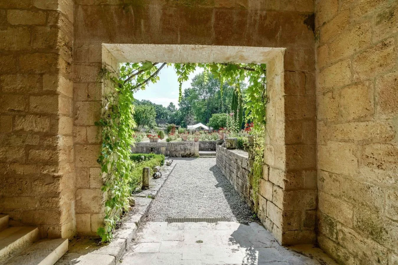 Stone archway opening onto a sunny landscaped garden with gravel paths, raised flower beds, and climbing vines.