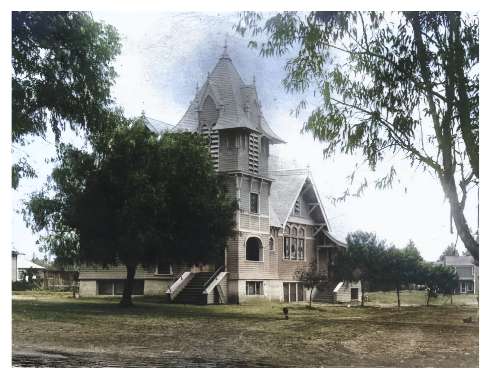 A large Victorian-style wooden house with a pointed tower, multiple gables, and stairs leading up to the front entrance, surrounded by trees and a grassy yard.