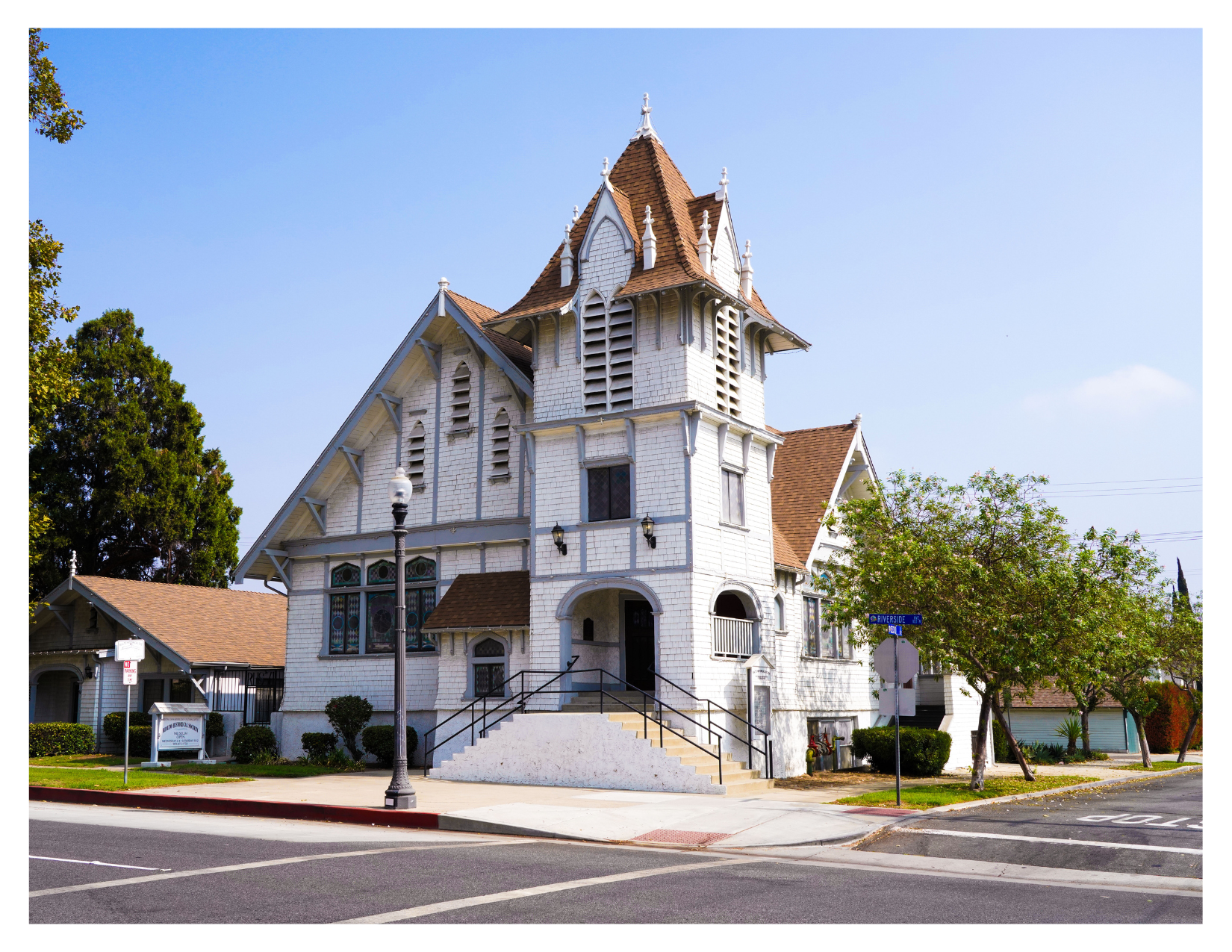 A historic white wooden church with a tall steeple and stained glass windows, located on a street corner with trees and street signs.