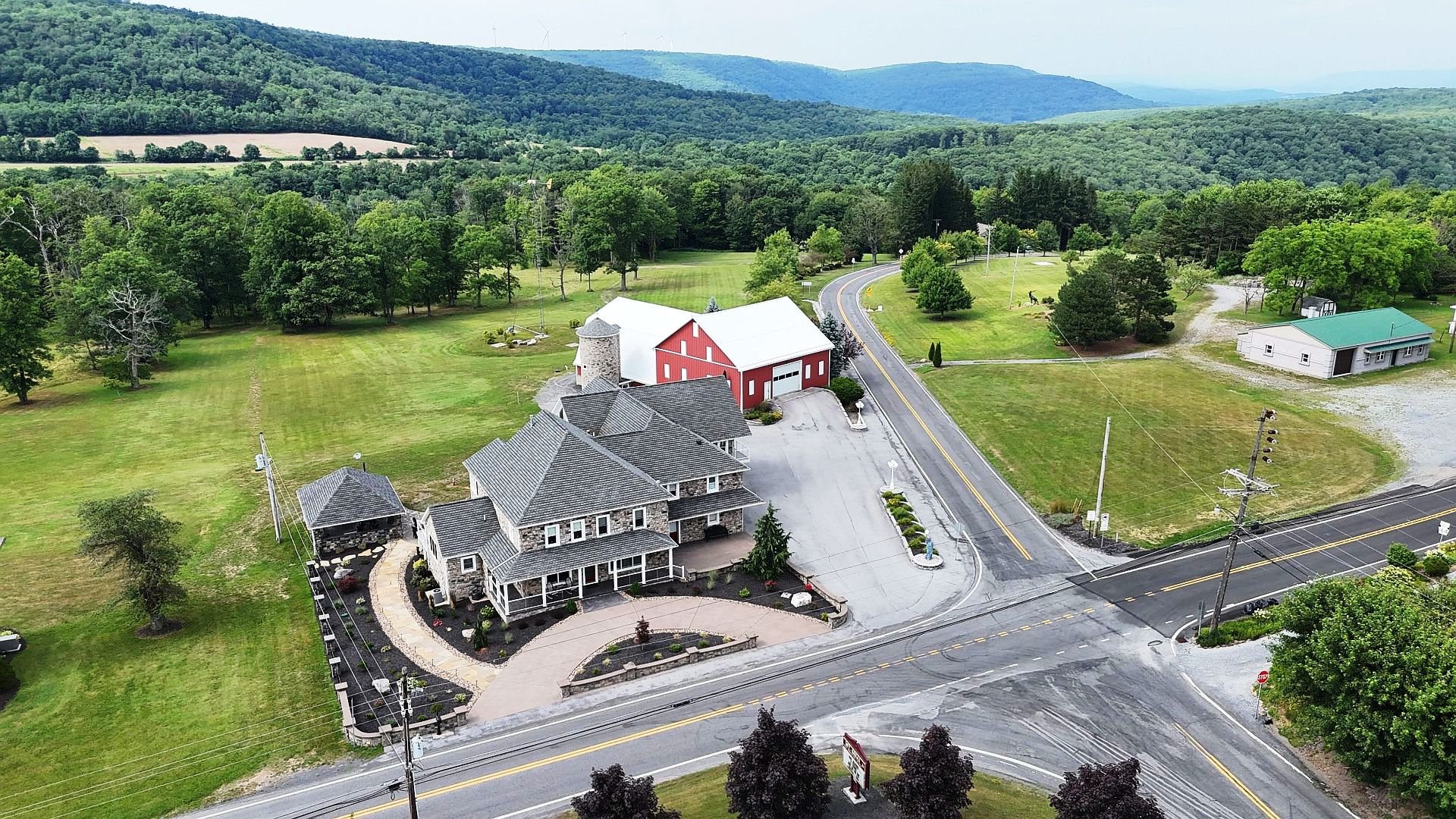An aerial farm photo of a rural property with a large stone house, a red barn, and surrounding green fields and trees. There are paved driveways, power lines, and a winding road in a mountainous landscape.
