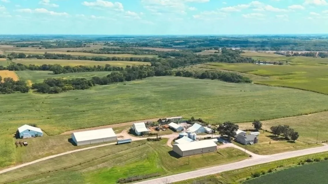 Aerial view of a farm with multiple white barns, silos, and out building surrounded by extensive green fields and farmland under a partly cloudy sky.