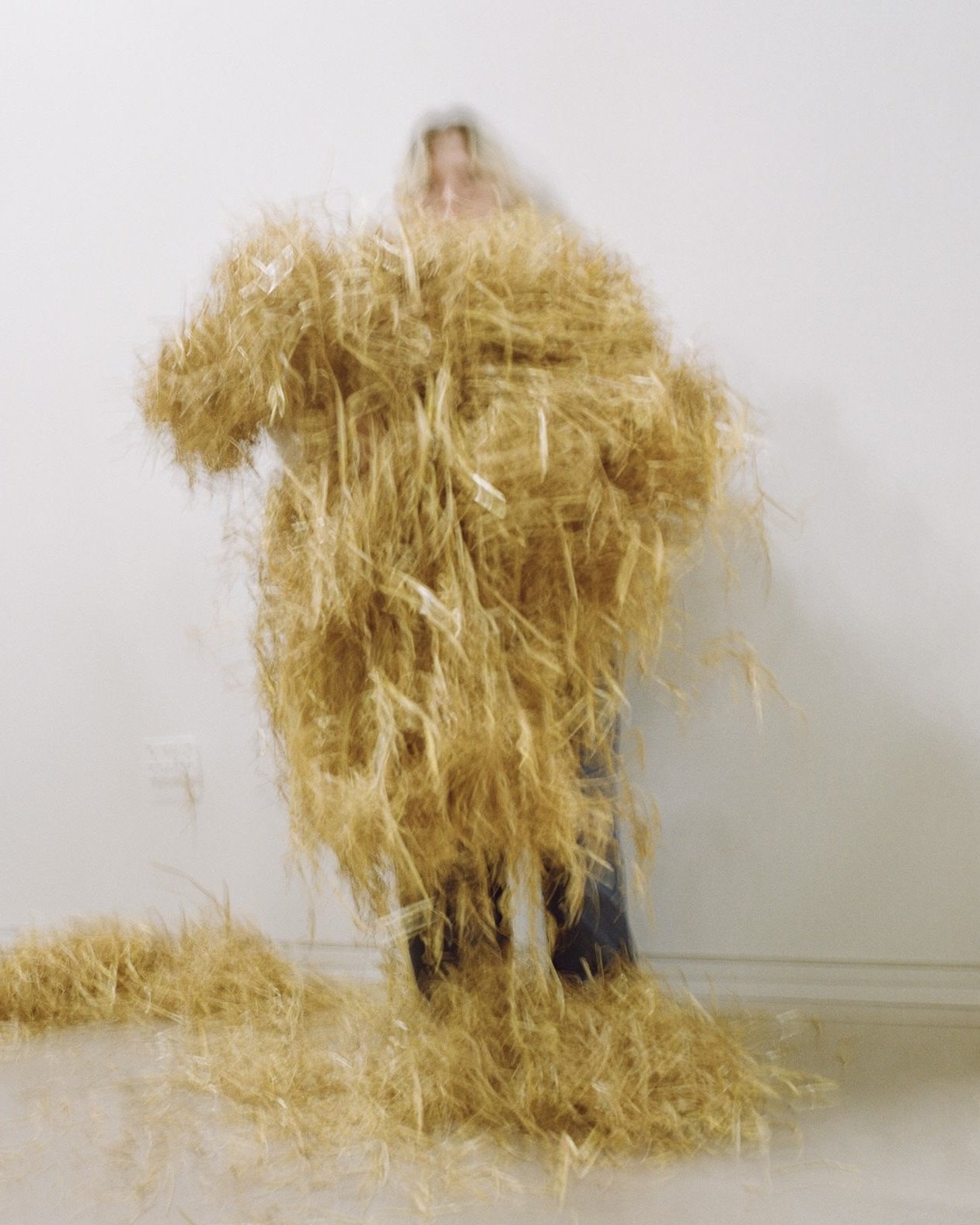 Cae McLaughlin standing behind a large pile of straw, with more straw on the floor in front of them, against a plain white wall.