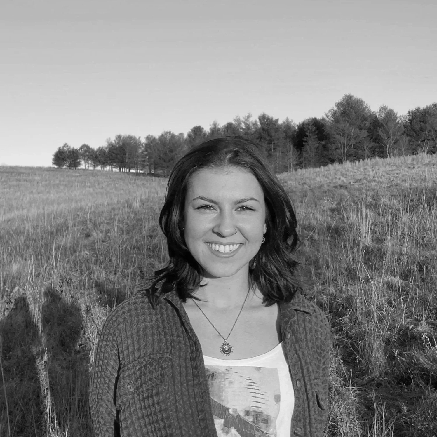 A woman smiling outdoors in a grassy field with trees in the background, captured in black and white.