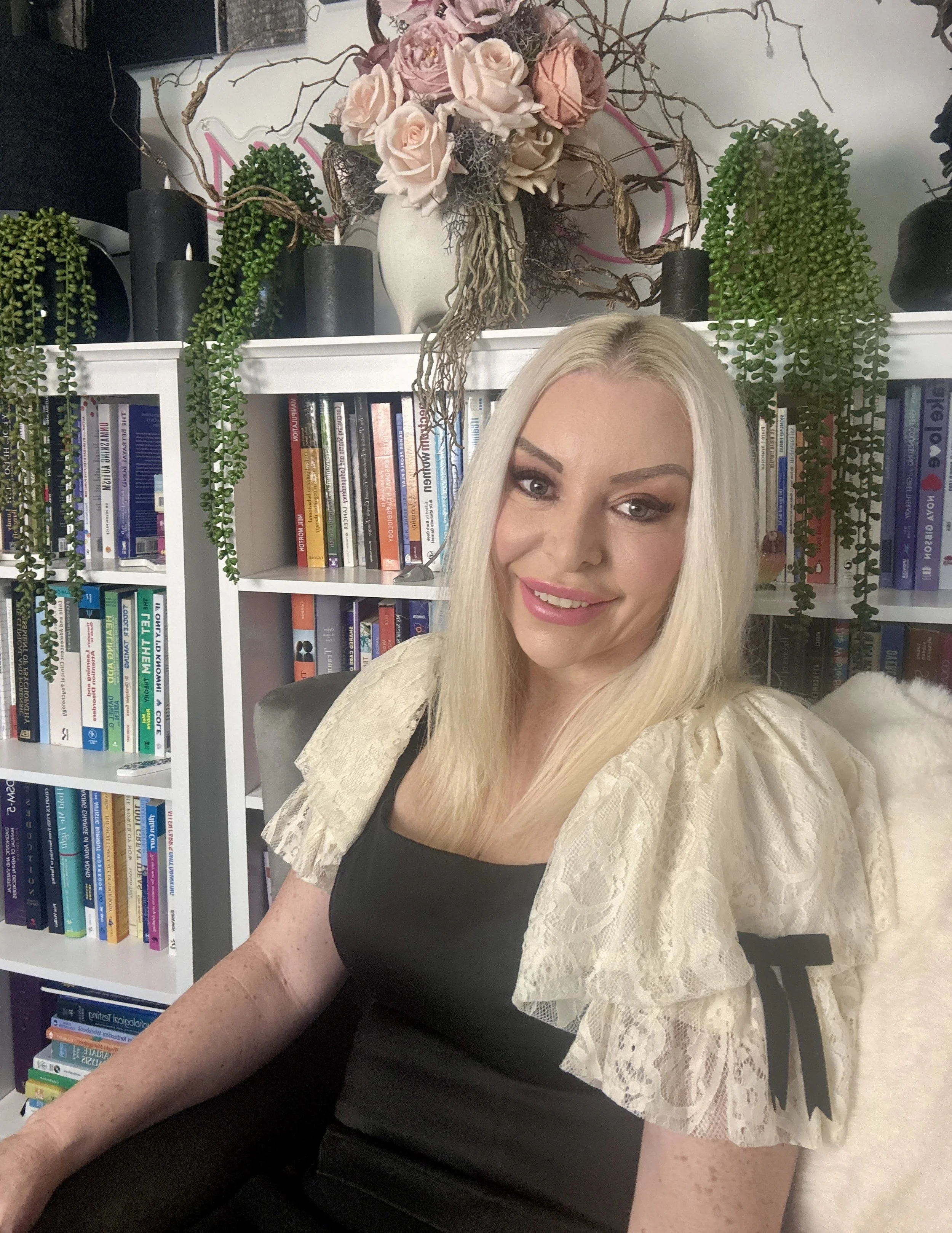 A woman with blonde hair sitting in front of a bookshelf filled with books, with a floral arrangement of pink and beige roses and green cascading plants on top of the bookshelf.