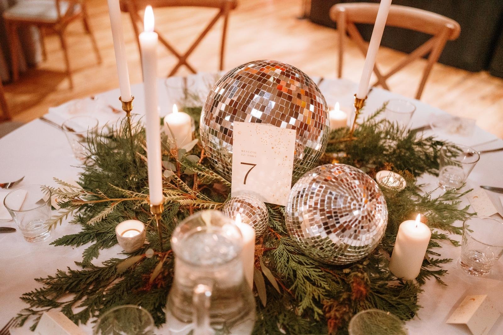 Decorative table centerpiece with three disco balls, candles, greenery, and a table number card.