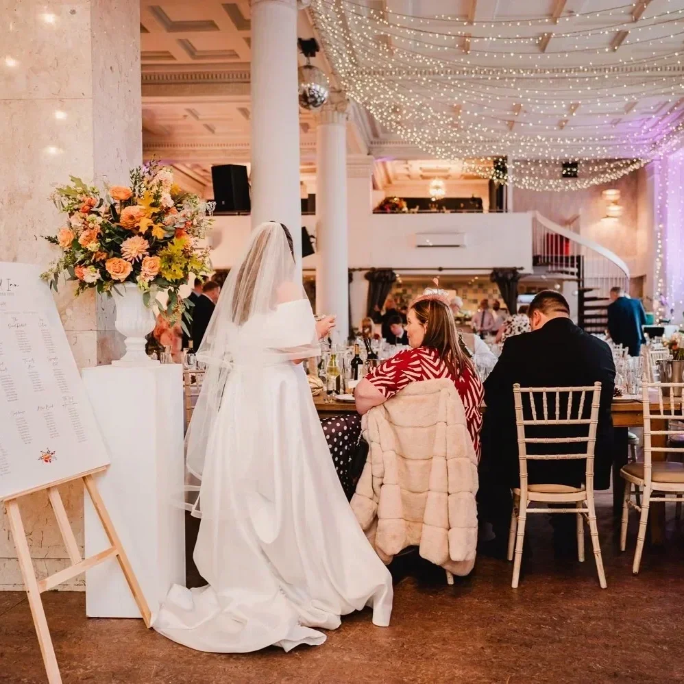 A bride in a white wedding gown and veil stands next to a table at a wedding reception. Guests are seated at the table, and the venue is decorated with string lights and floral arrangements.
